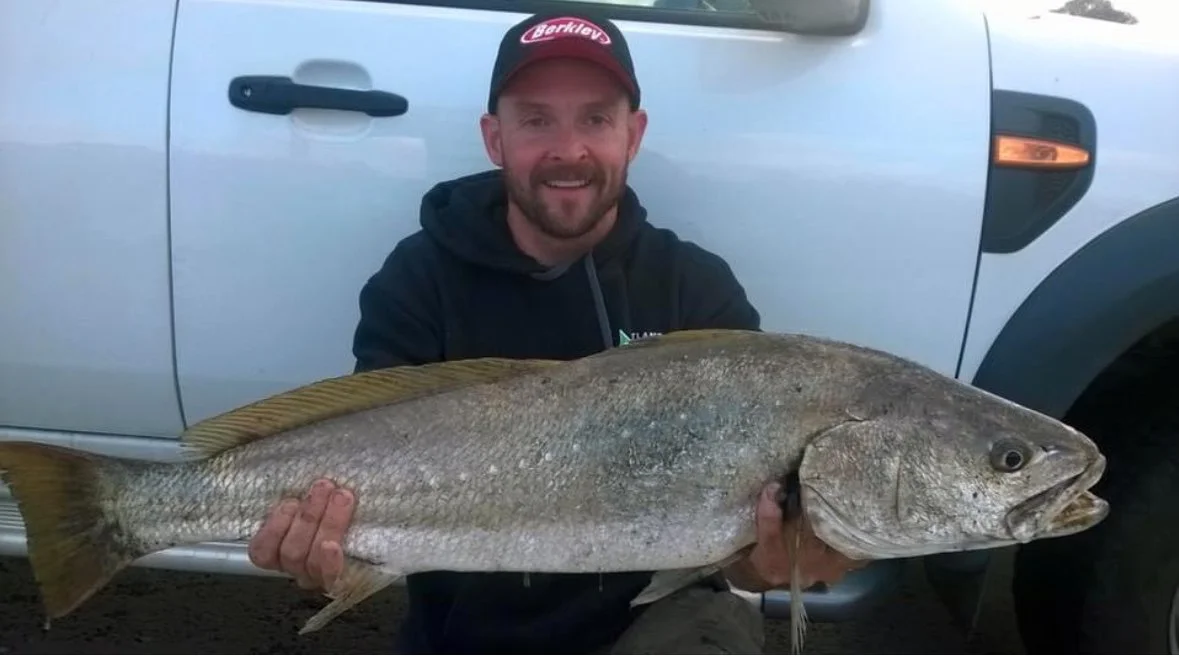 Man in a dark hoodie and red cap smiling while holding a large fish in front of a white vehicle.