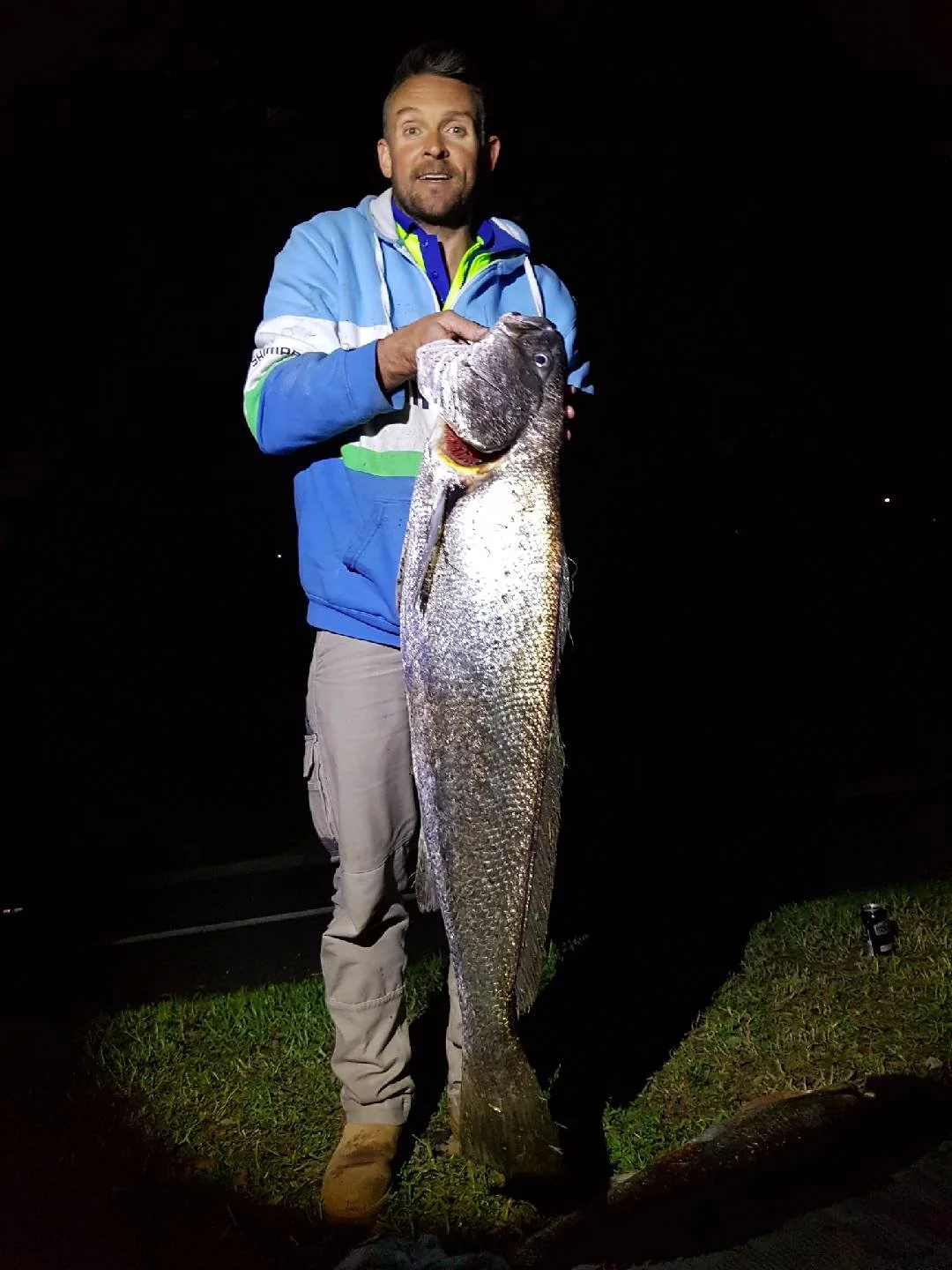 A man in a blue jacket and beige pants holding a large fish at night.