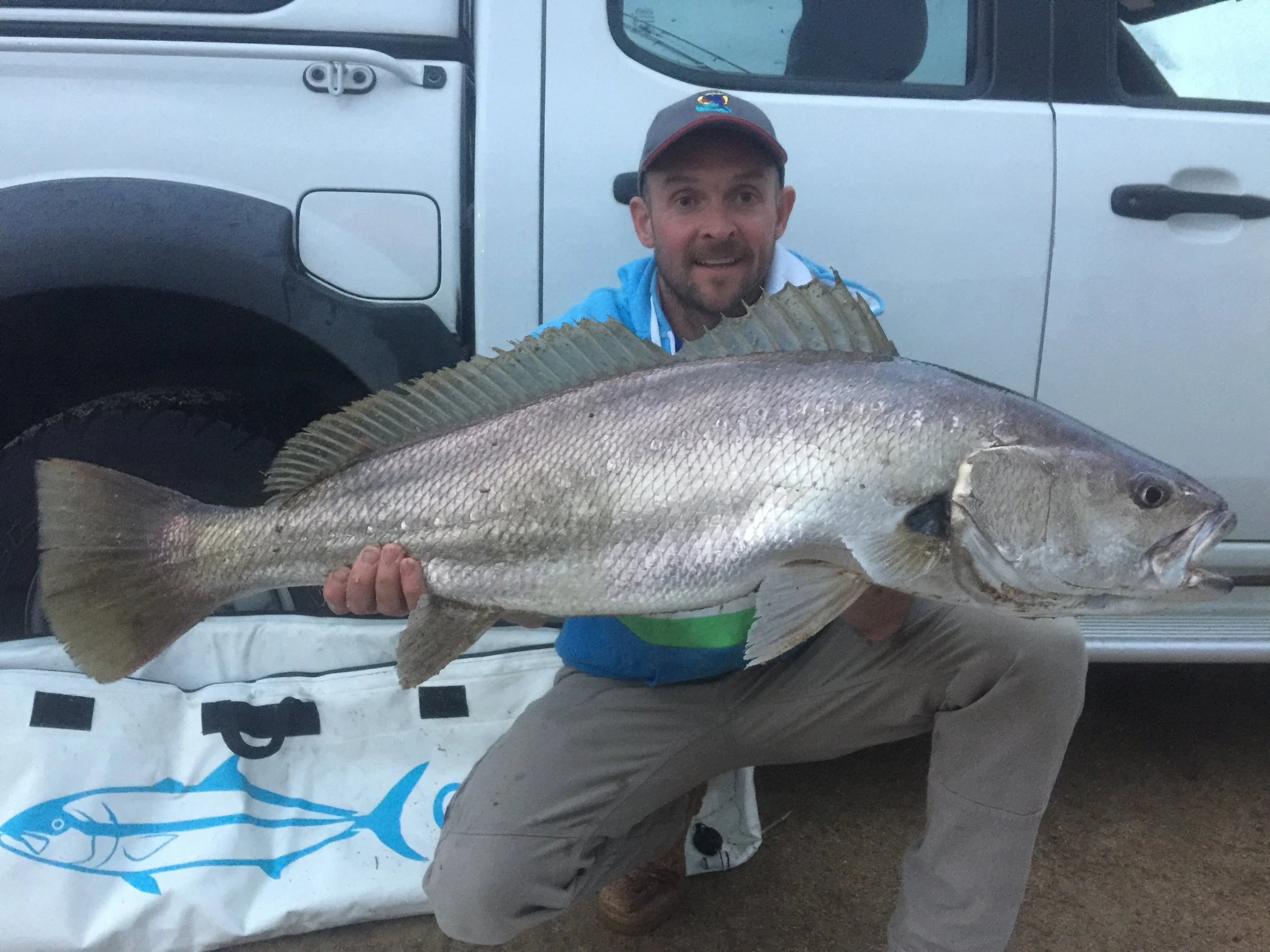 A man wearing a baseball cap and a blue jacket holding a large fish in front of a white vehicle with black accents.