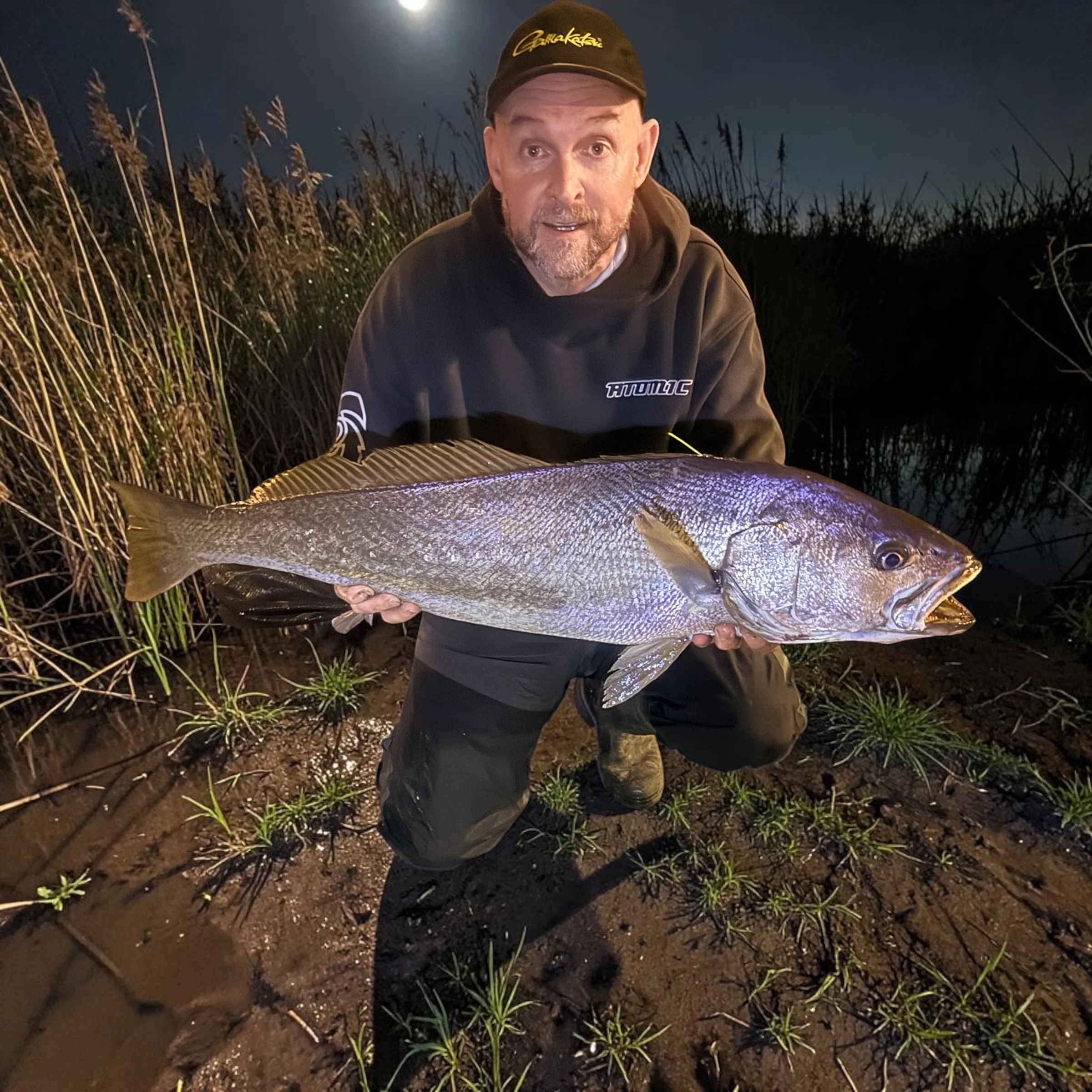 A man with a beard wearing a black hoodie and a black cap, crouching in a muddy field at night, holding a large fish with shiny scales.