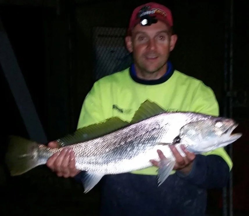 Man holding a large fish outdoors at night, wearing a neon yellow jacket and a red cap.