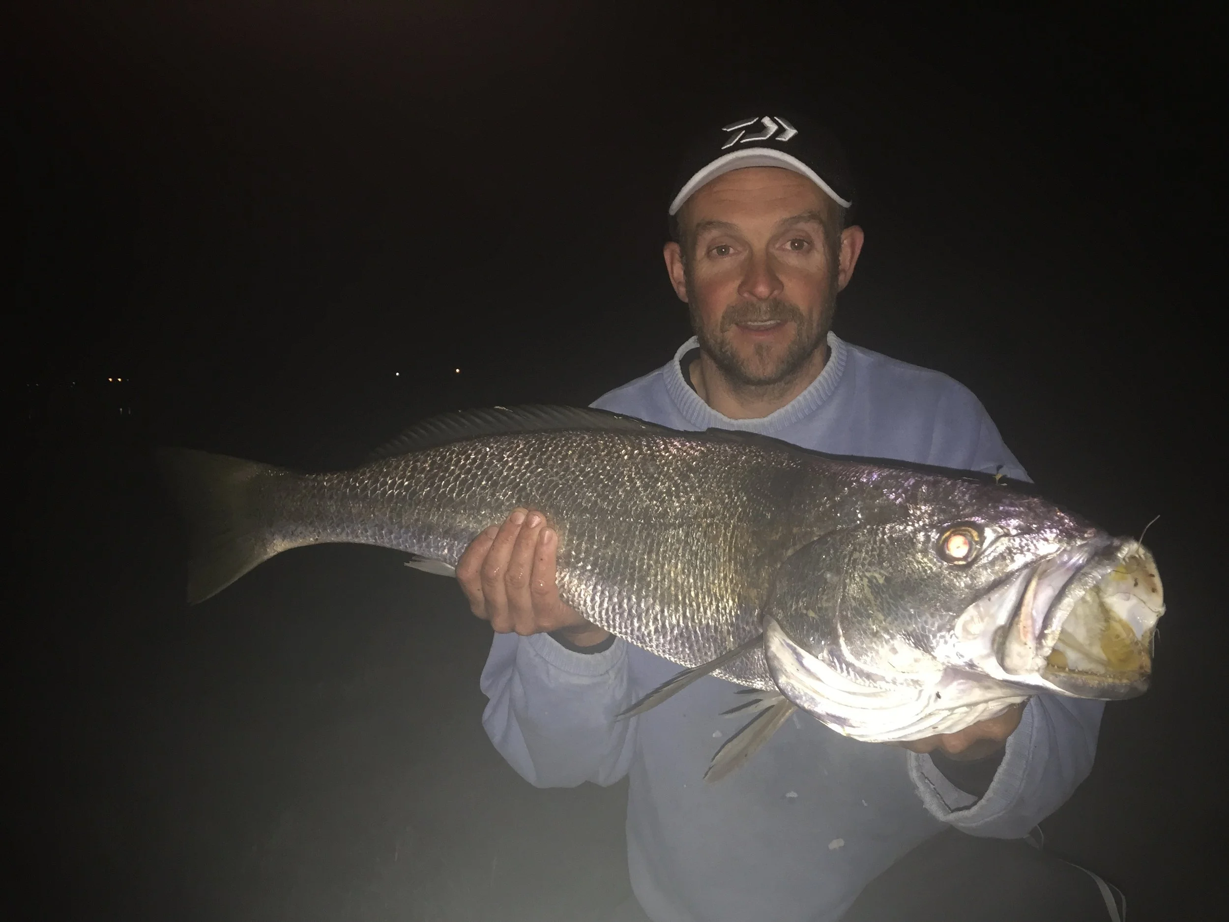 A man at night holding a large fish, wearing a white cap with a black logo, and a light-colored sweatshirt.