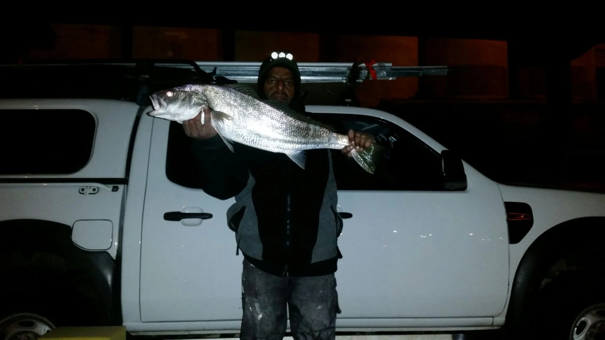 A man holding a large fish in front of a white truck at night.
