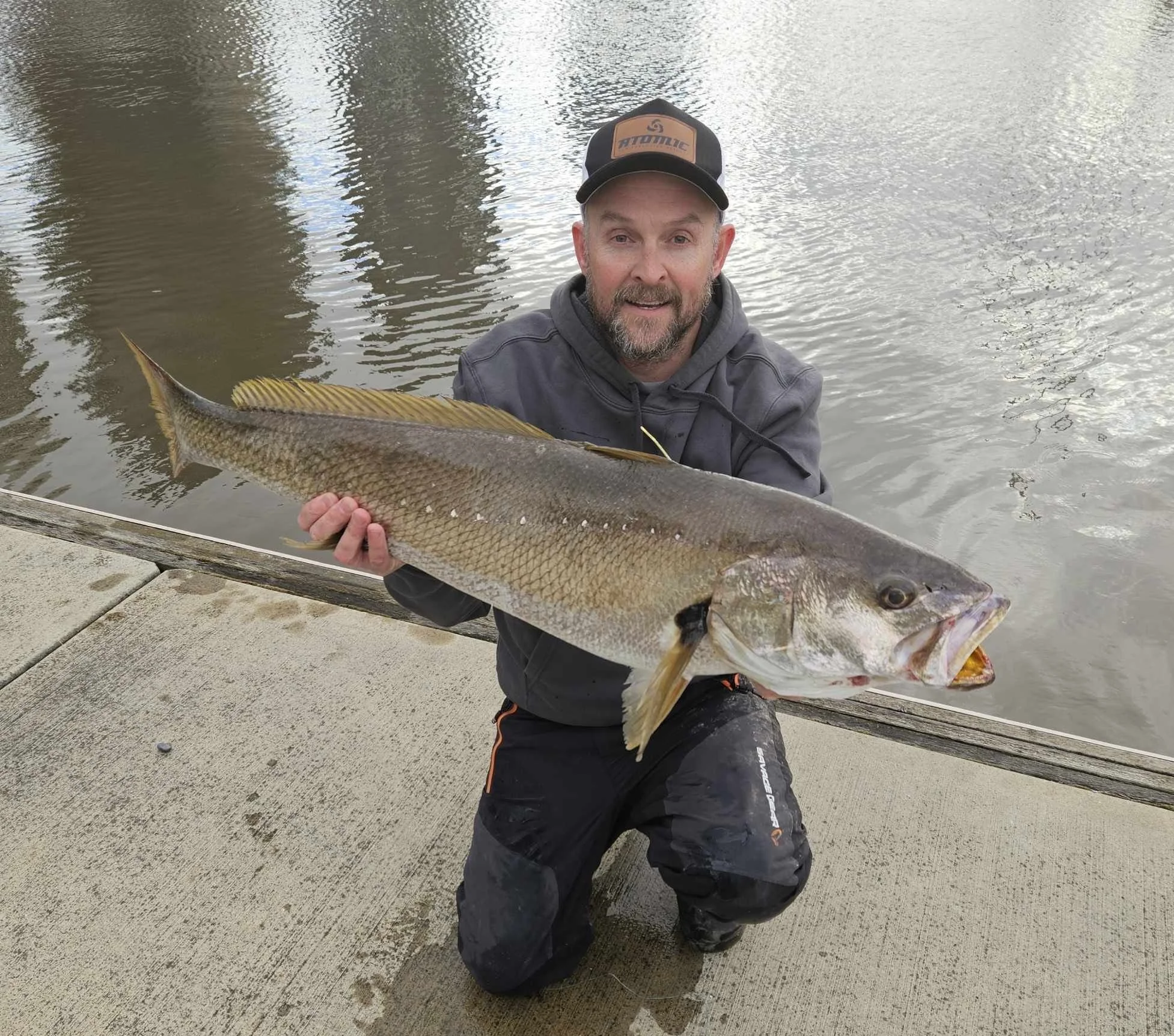 Man in black pants and gray jacket holding a large fish near a body of water with overcast sky.