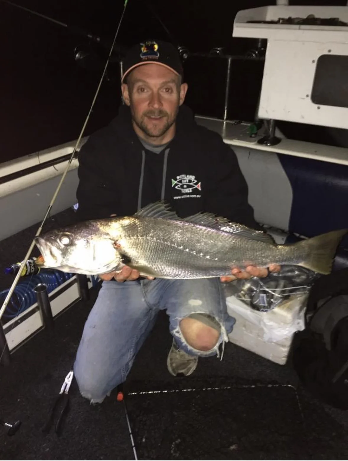 A man in a black hoodie and ripped jeans kneeling inside a boat at night, holding a large fish with both hands. The boat has fishing equipment and a cooler, with a white structure in the background.