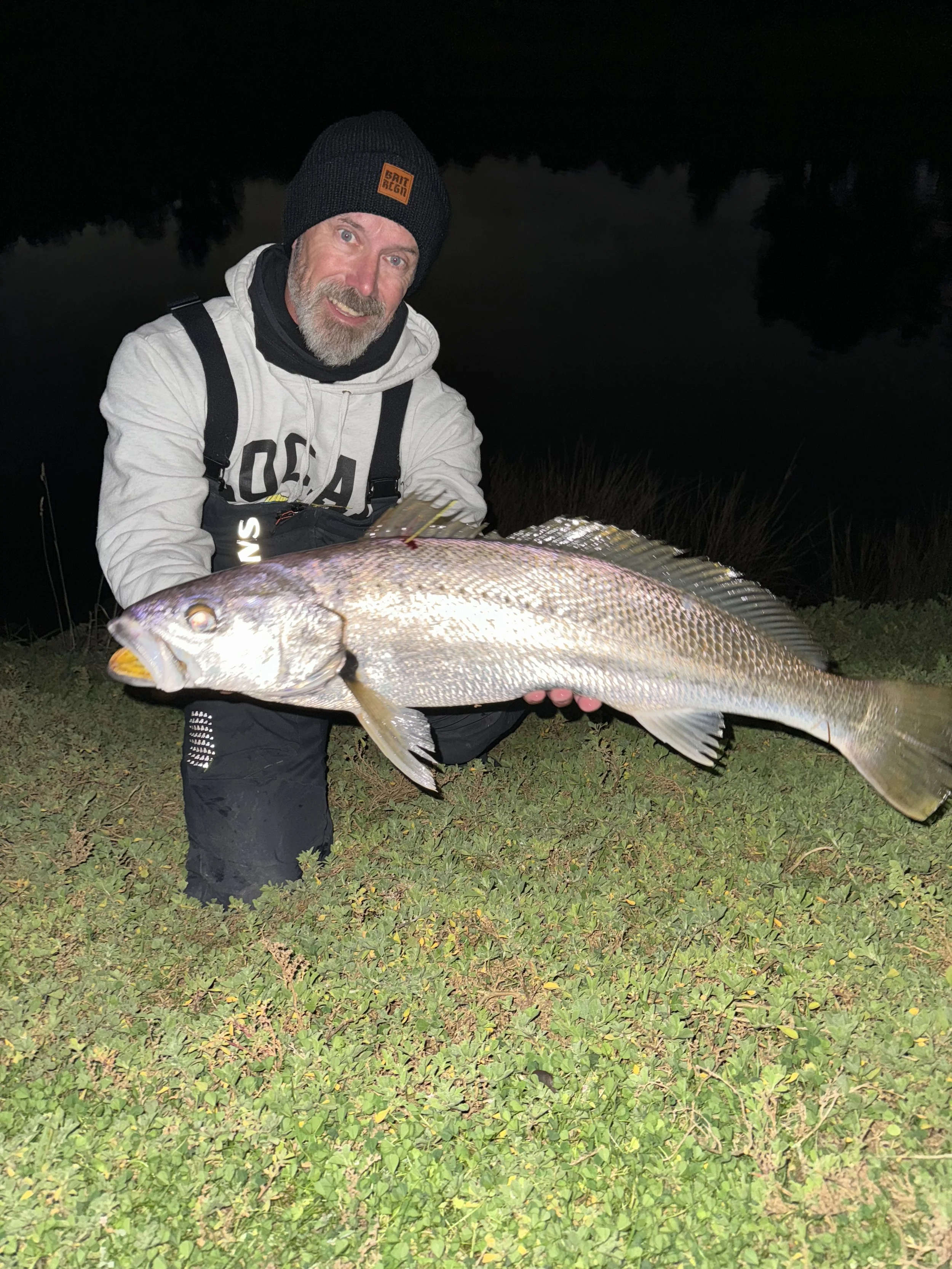 A man with a beard in a white hoodie and black beanie holding a large fish outdoors at night, with grass underfoot and dark water and trees in the background.