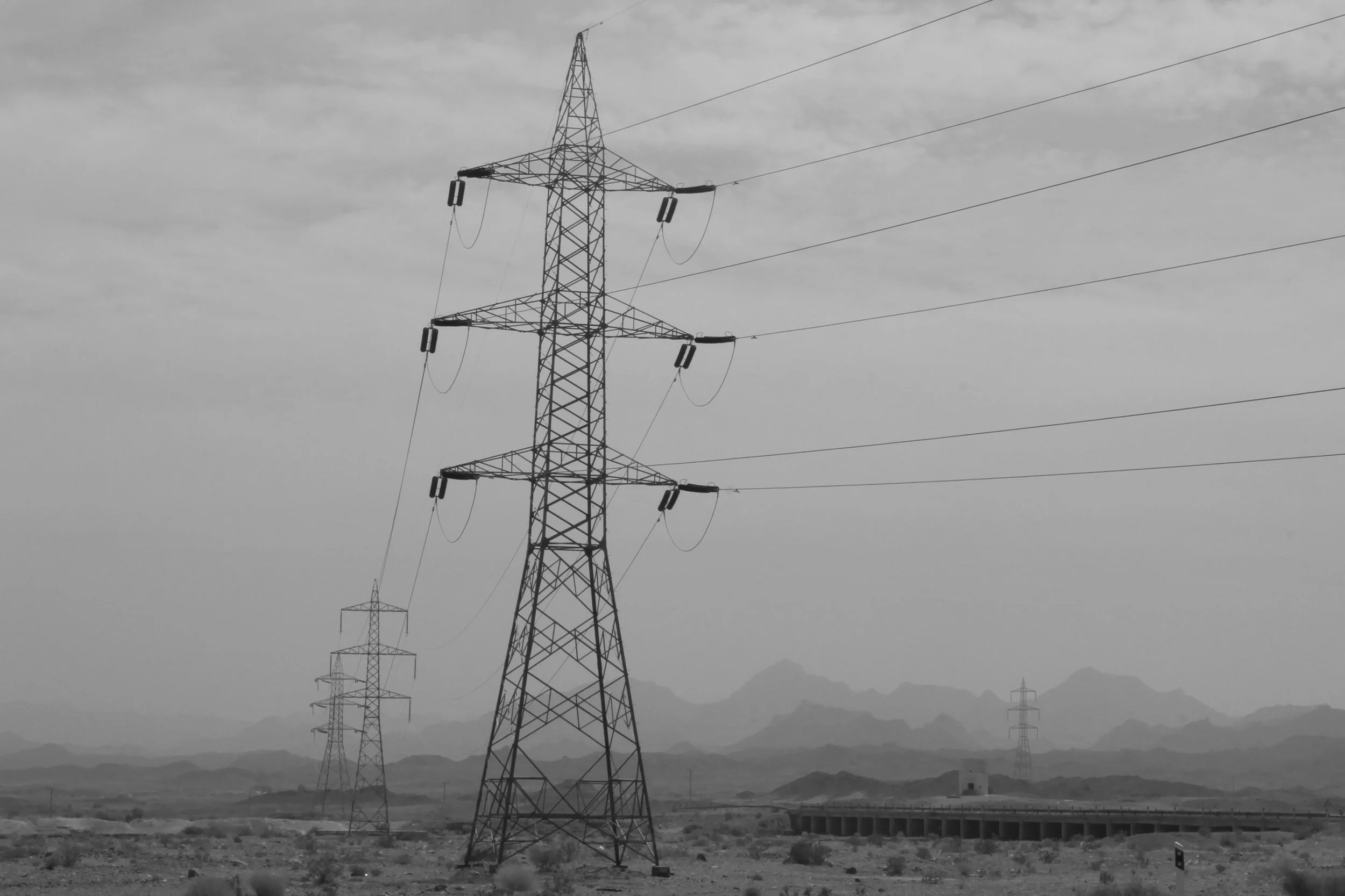 The structure dominates the landscape, yet what it carries remains invisible. Power reveals itself only through its effects, never through its own presence. The tower stands as a monument to an absence that organizes the visible world. Between earth 