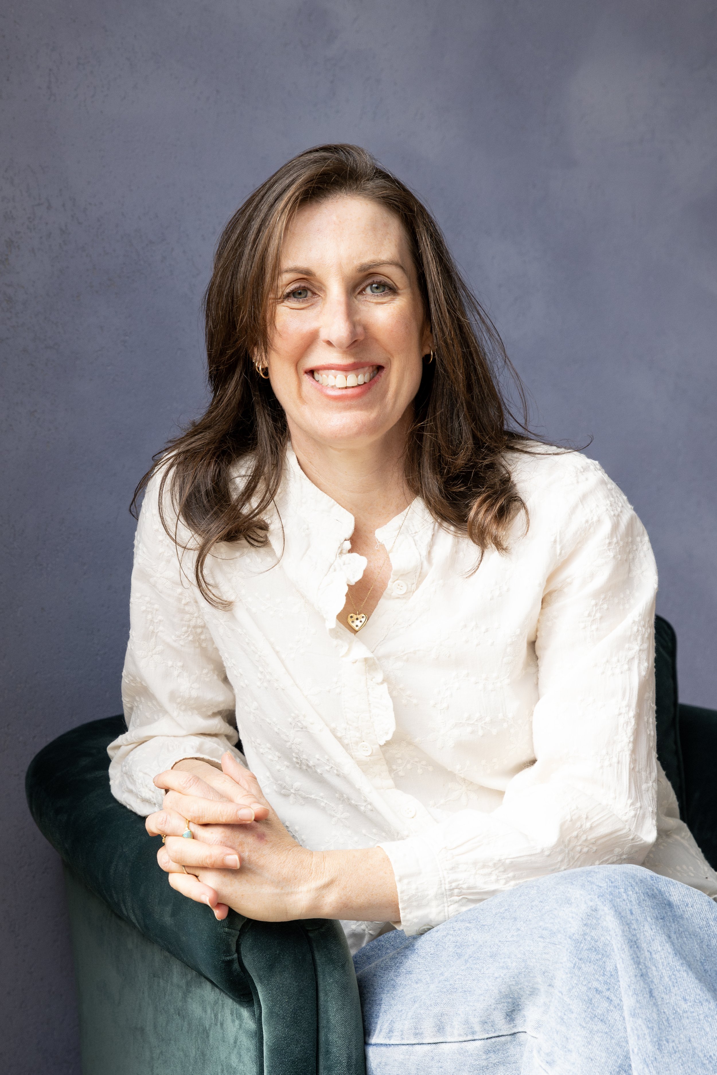 A woman with shoulder-length brown hair, wearing a cream-colored shirt, sitting on a dark green chair against a plain gray background, smiling at the camera.