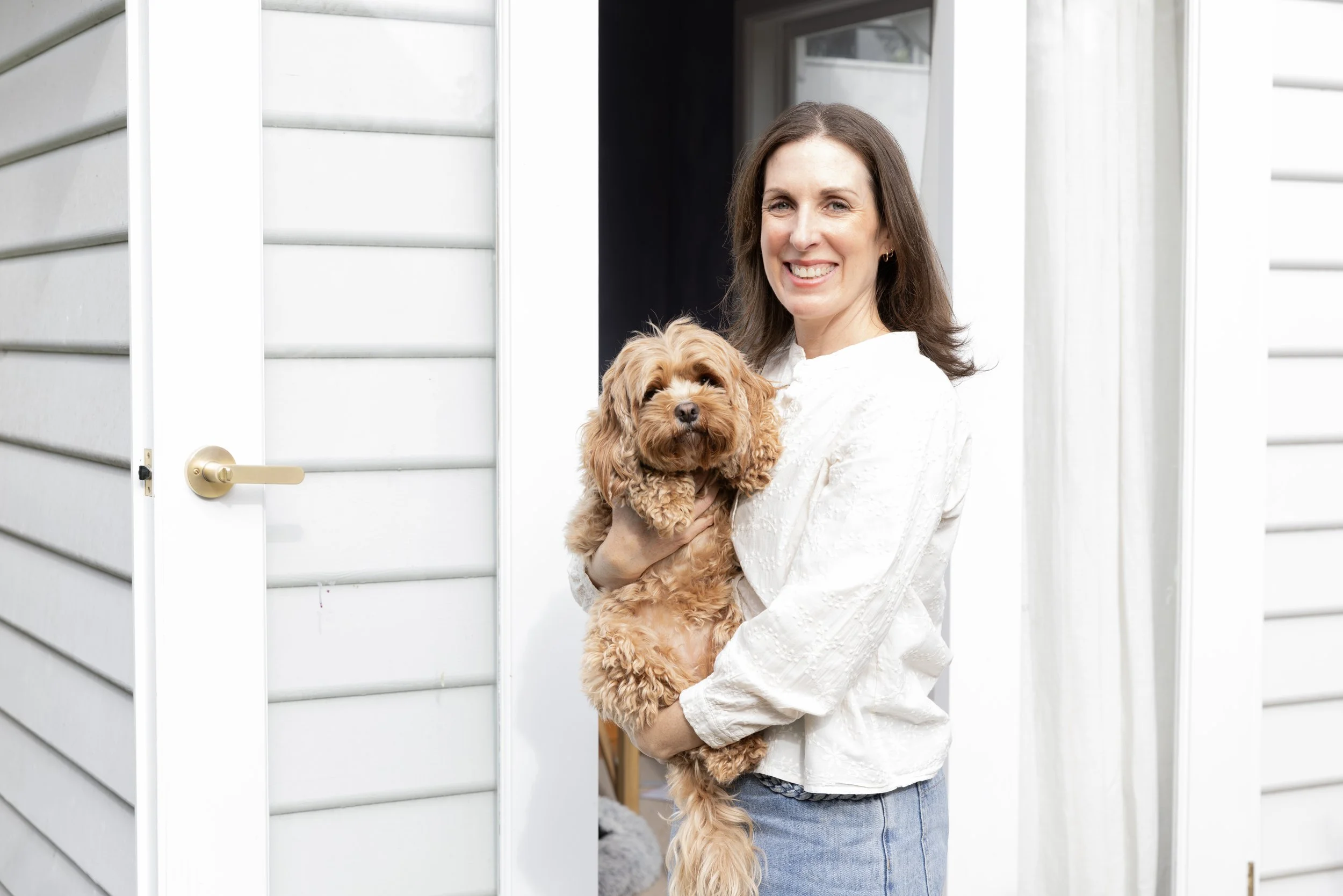 A woman smiling while holding a small, brown, fluffy dog. She is standing in a doorway with white siding, and there is an indoor space visible behind her.