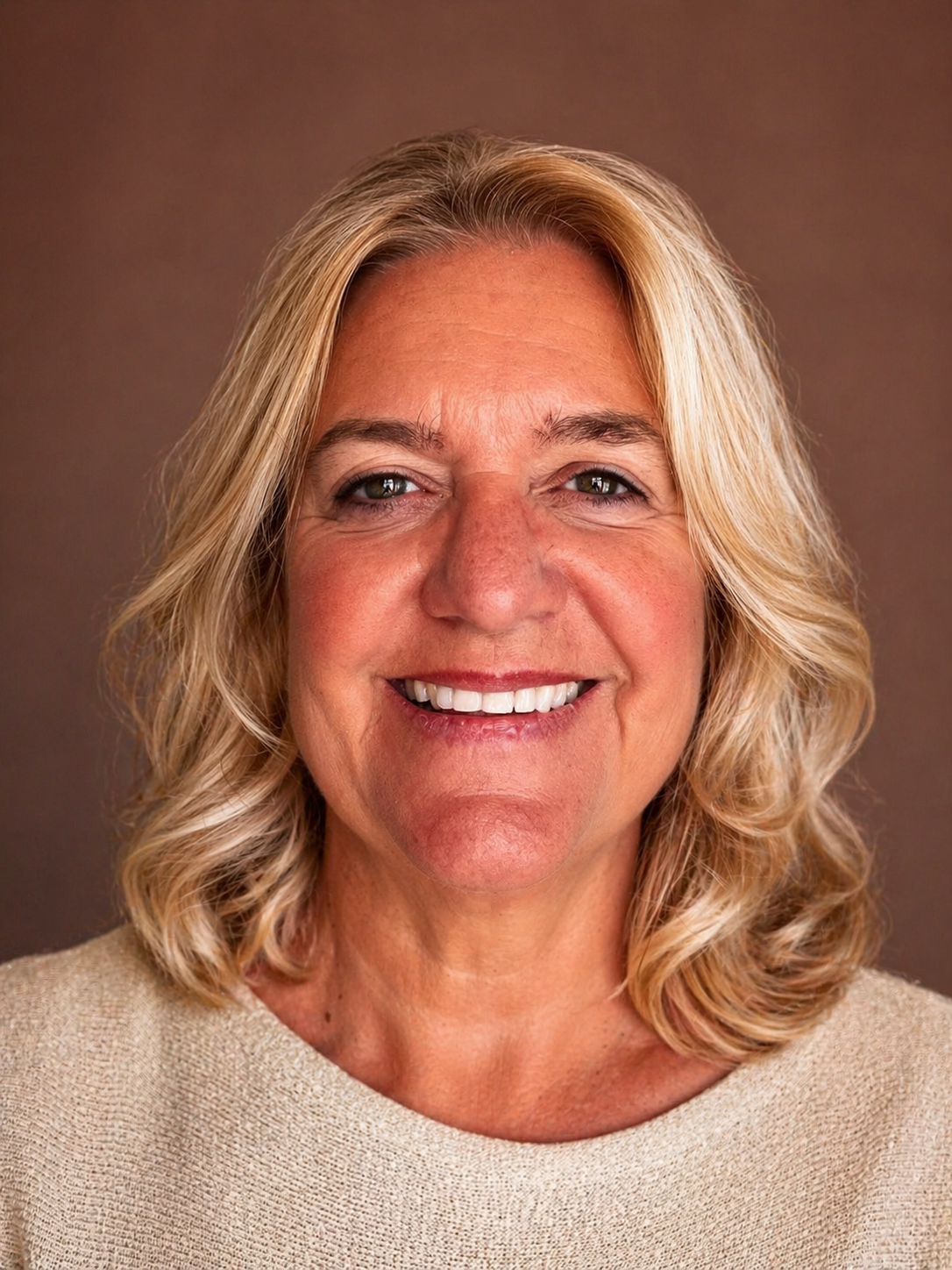 Close-up portrait of a middle-aged woman with blonde, wavy hair, smiling and wearing a beige top against a plain brown background.