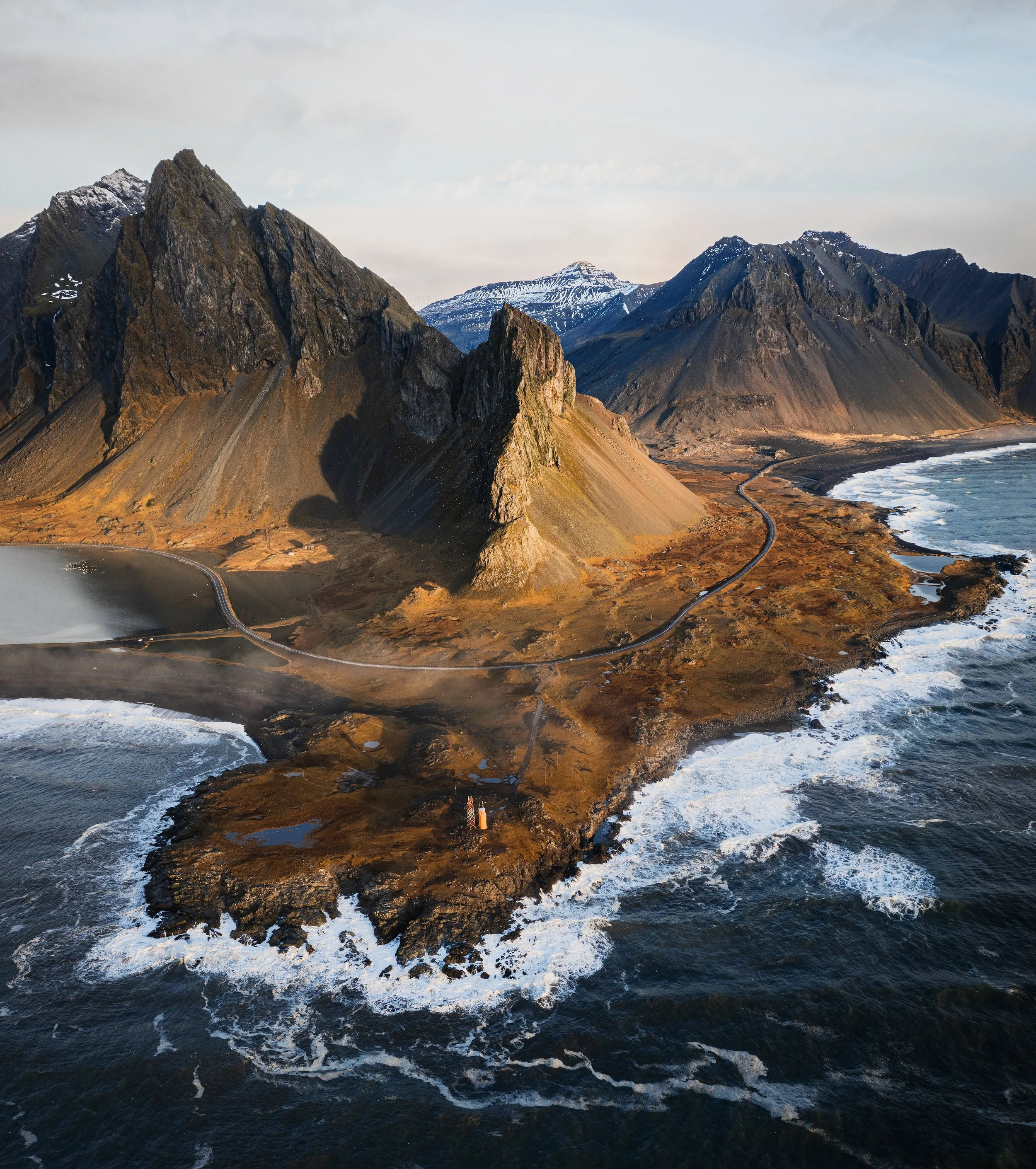 A rugged coastal landscape featuring tall, jagged mountains with patches of snow. The foreground shows dark rocky shoreline with waves crashing against it. A narrow road winds along the shoreline, and the sky is partly cloudy.