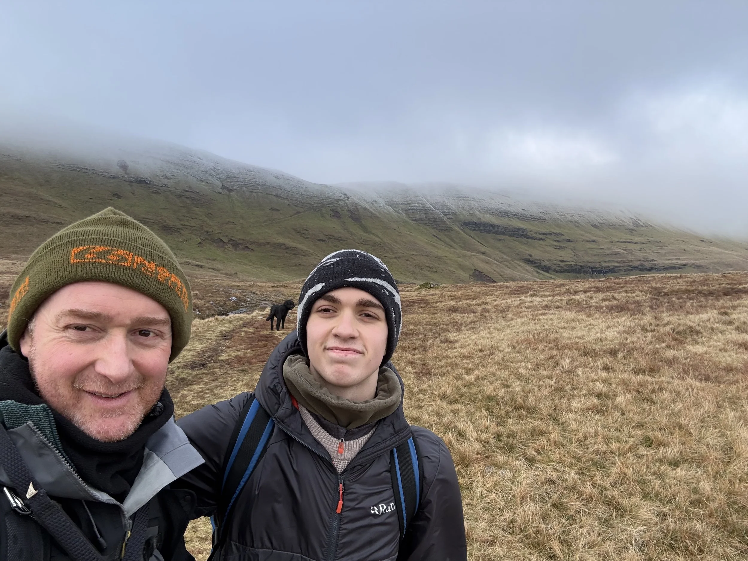 Two people taking a selfie in a natural landscape with hills or mountains, some snow on the peaks, grassy terrain, and clouds or mist in the sky.