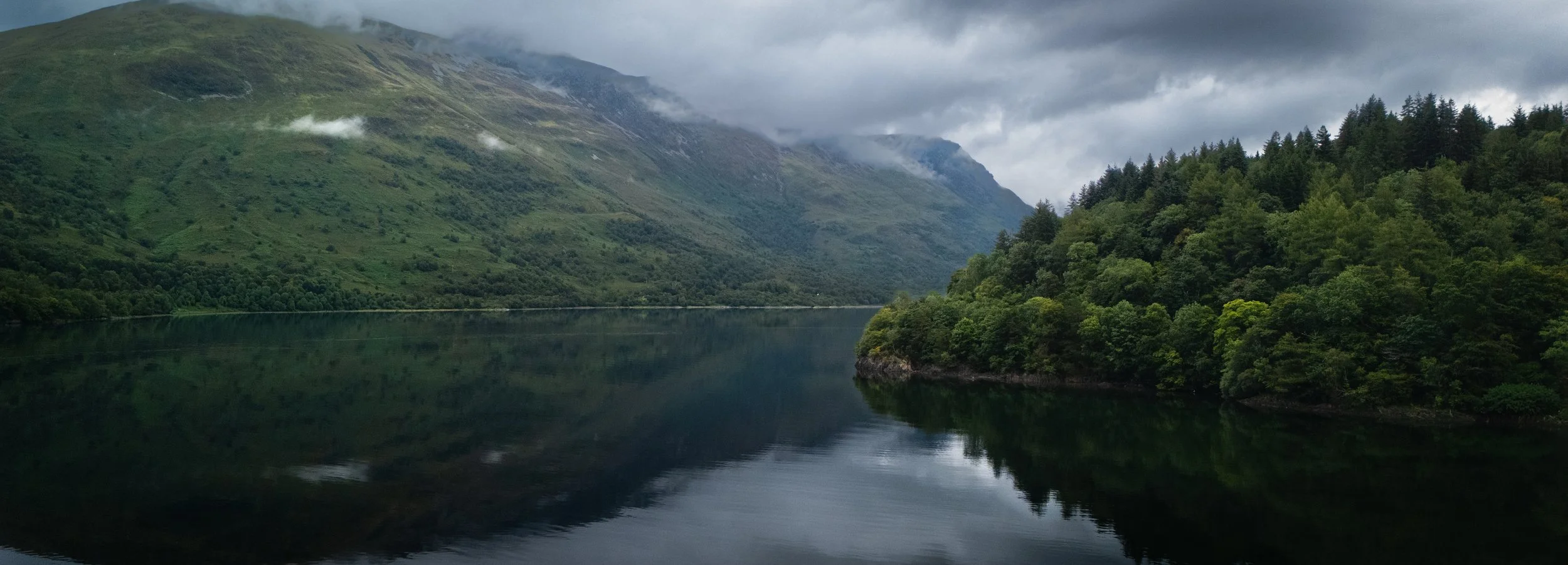 Scenic view of a calm river surrounded by lush green mountains with clouds overhead.