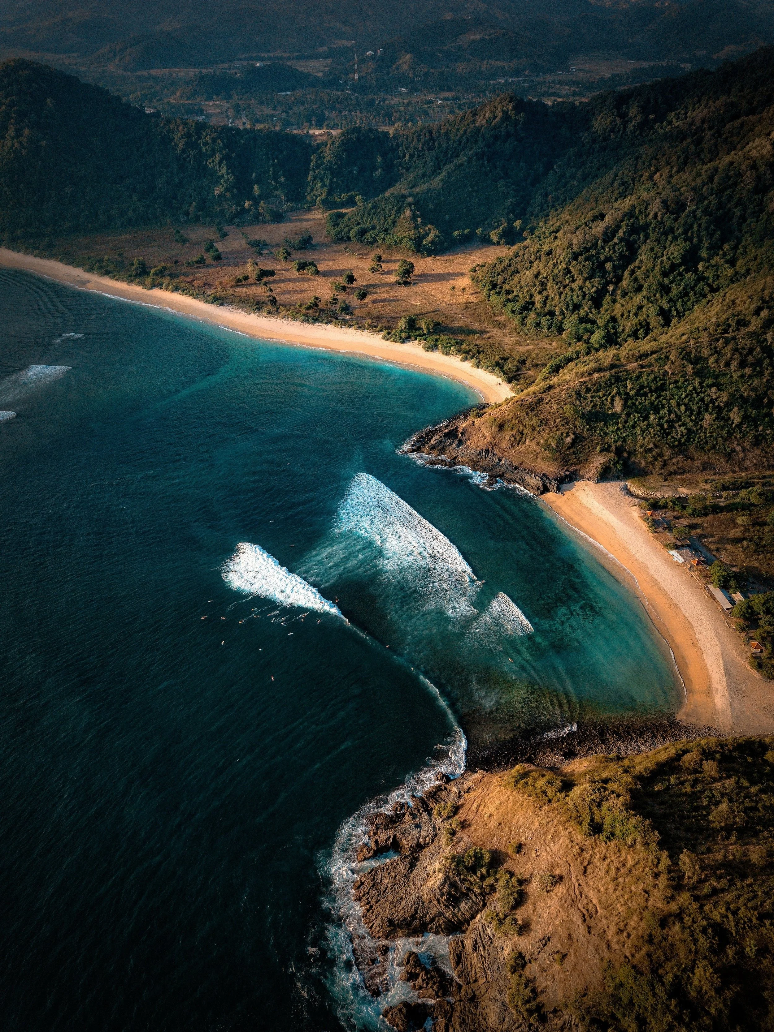 Aerial view of a coastline featuring clear blue ocean waves crashing against a rocky shore, with a sandy beach and lush green hills in the background.