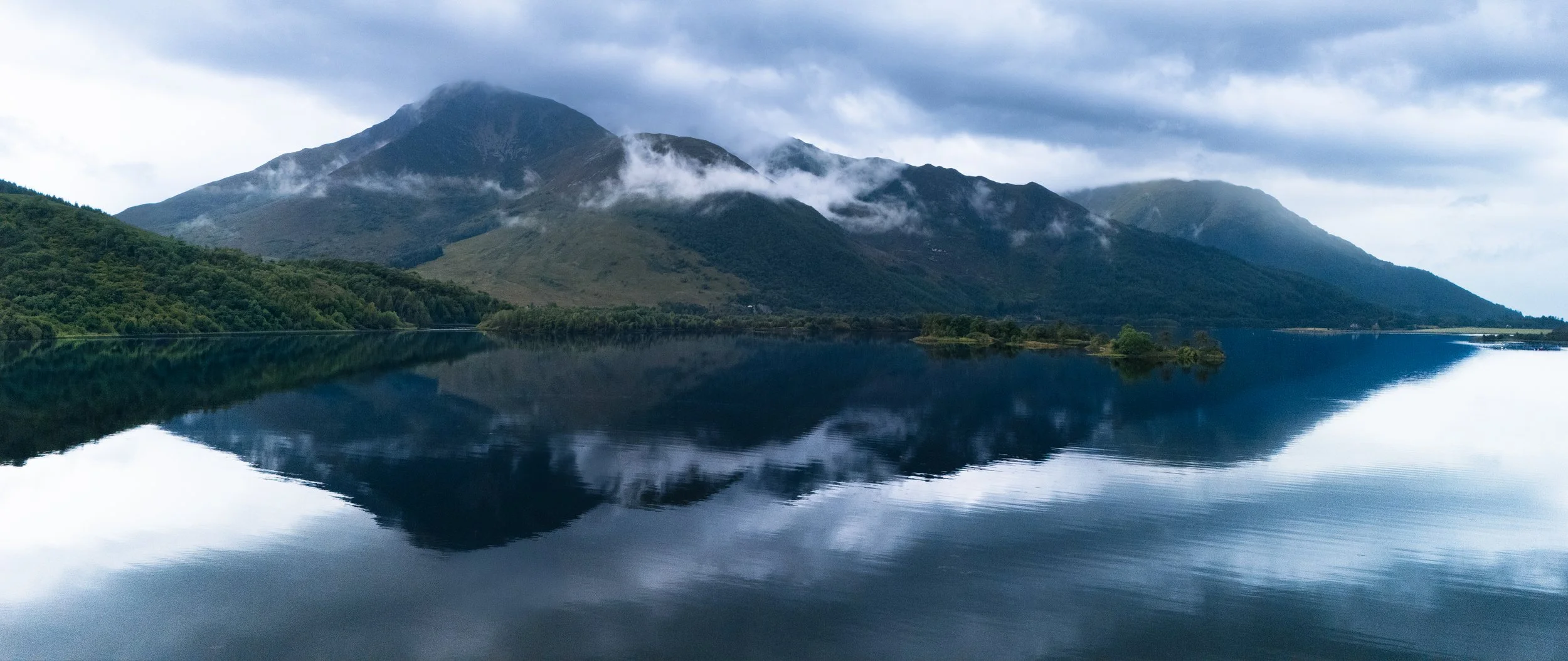 Mountains with clouds reflected in a calm lake beneath a cloudy sky.