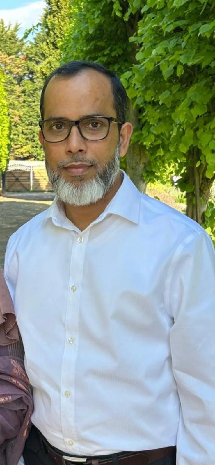 A man with glasses and a gray beard standing outdoors in a green garden, wearing a white button-down shirt.