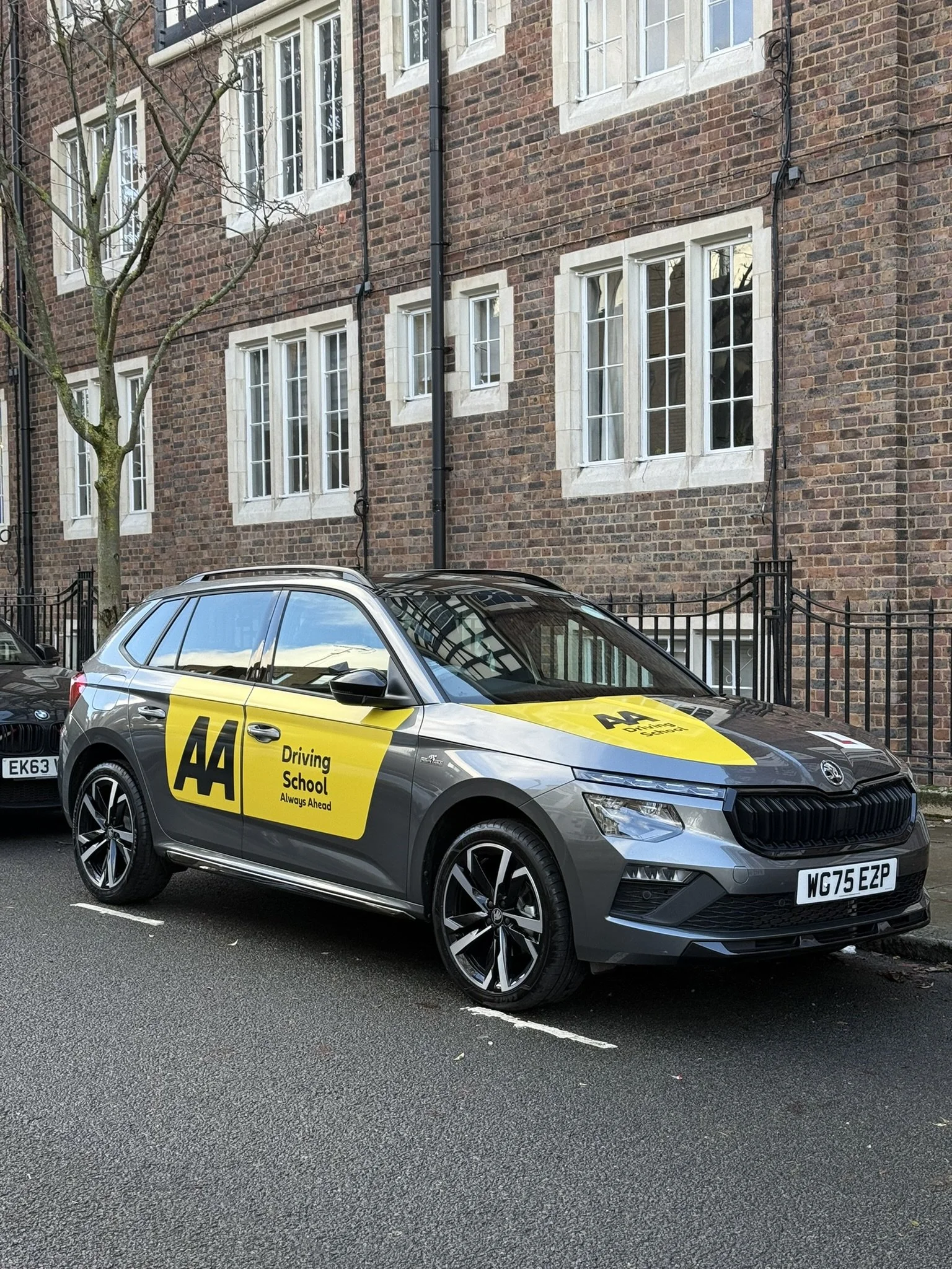 A silver Skoda car with AA Driving School livery parked on a street in front of a brick building with multiple windows.