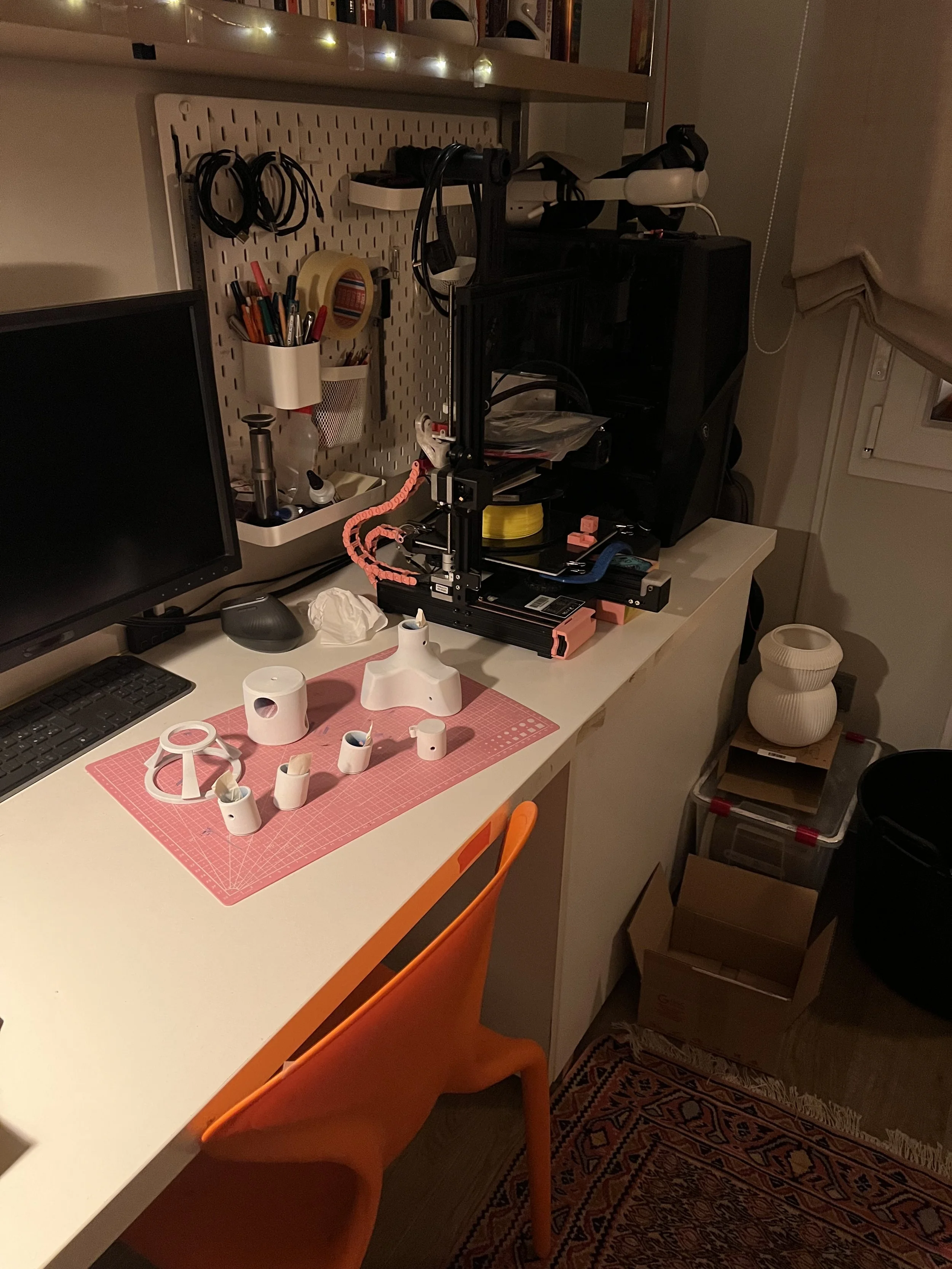 A cluttered workspace with a computer monitor, keyboard, mouse, and various tools and materials for 3D printing or crafting on the desk, including a 3D printer, filament spool, and small assembled plastic components.