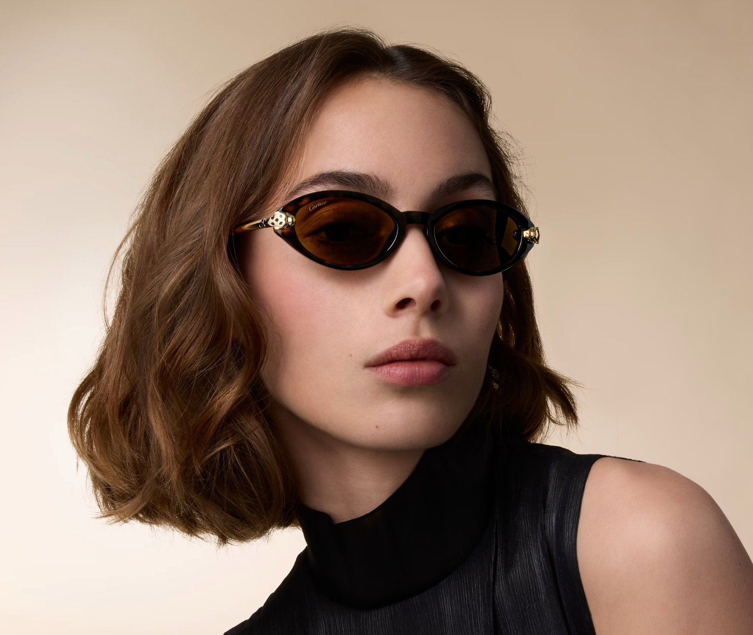 A woman with wavy brown hair wearing black and gold sunglasses and a black sleeveless top, posing against a neutral background.