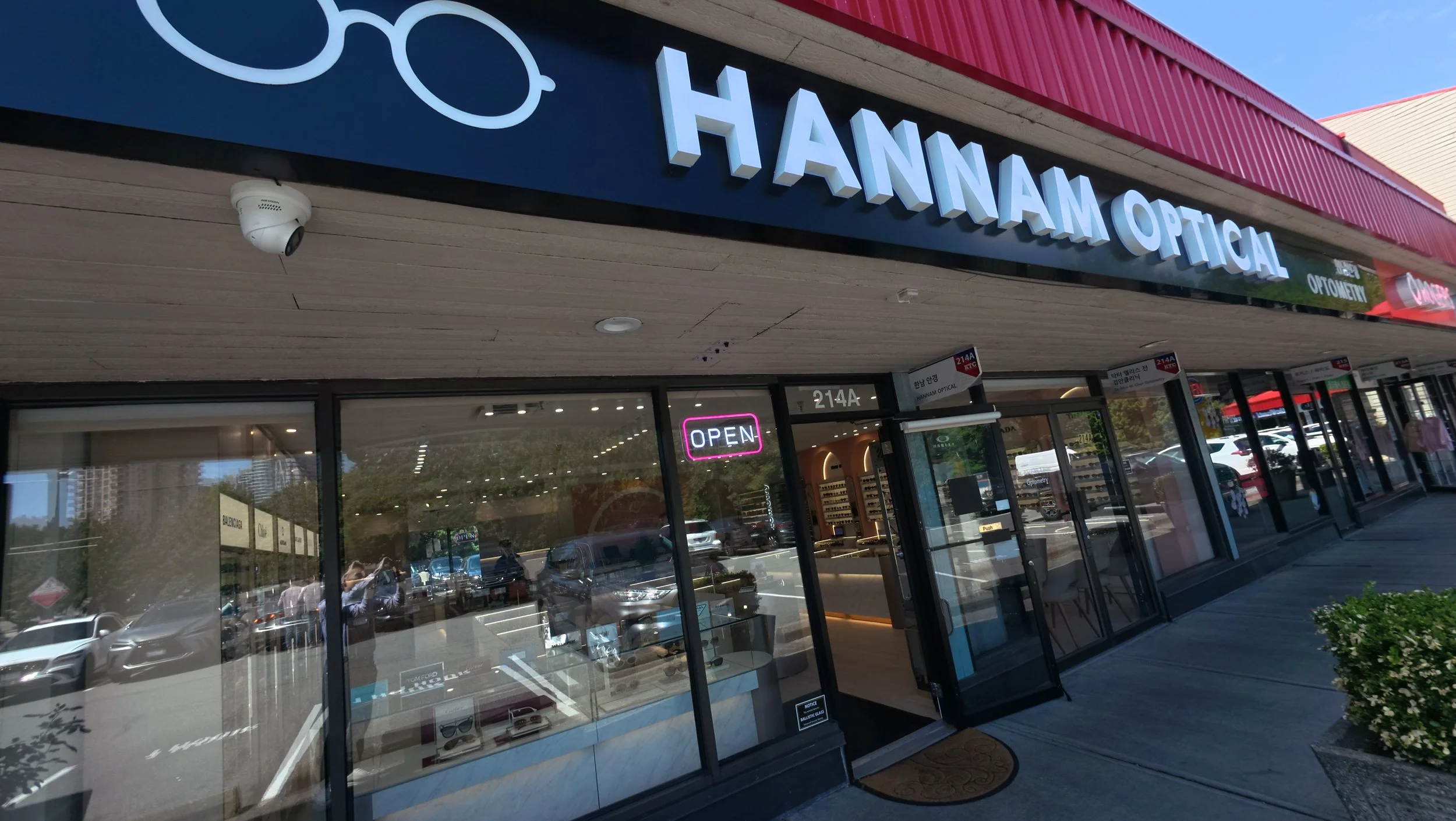 Exterior view of Hannam Optical store with large sign, glass doors showing interior, and a pink illuminated 'Open' sign. Reflections of parked cars and trees can be seen in the glass.