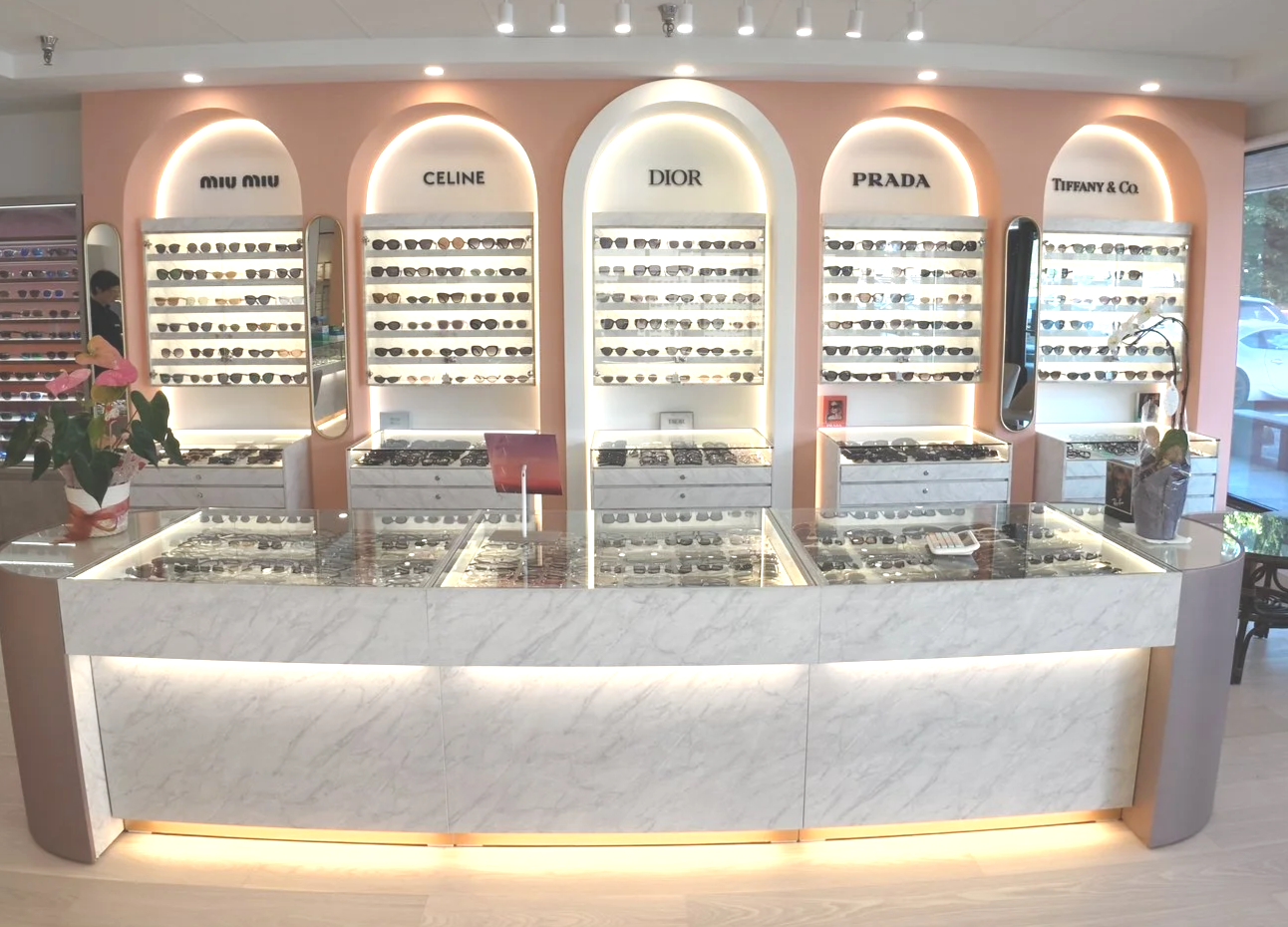 Display of designer sunglasses from brands Miu Miu, Celine, Dior, Prada, and Tiffany & Co. in a stylish store with marble counter, pink and white decor, and bright lighting.