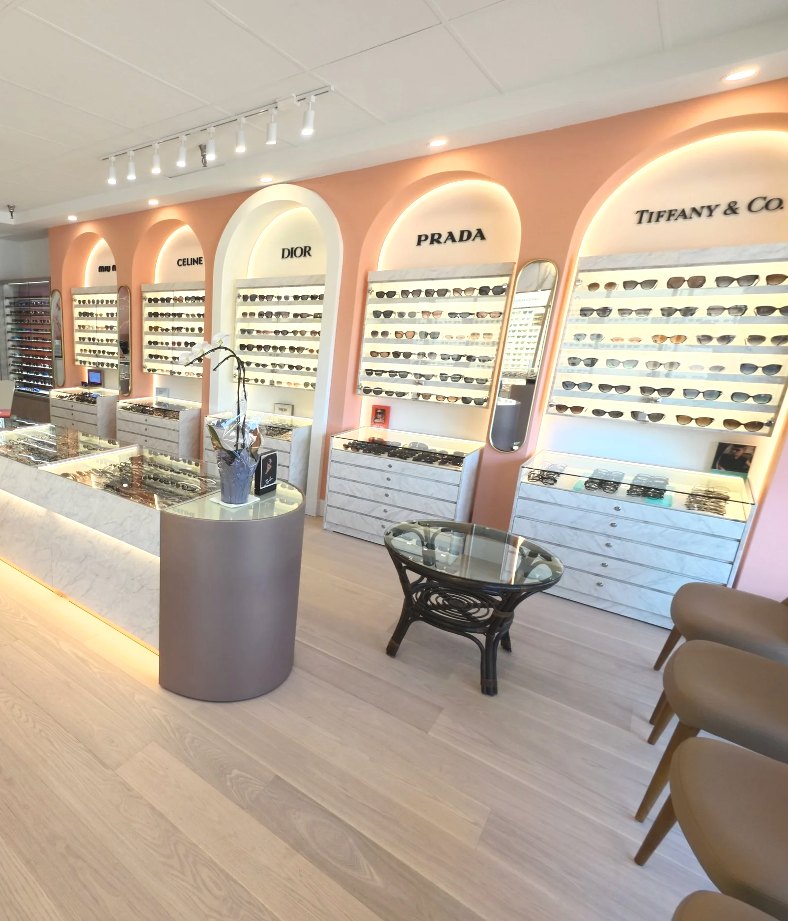 Interior of a sunglasses store with display shelves named after high-end brands like Celine, Dior, Prada, and Tiffany & Co. There are chairs for customers and lighting highlighting the sunglasses.
