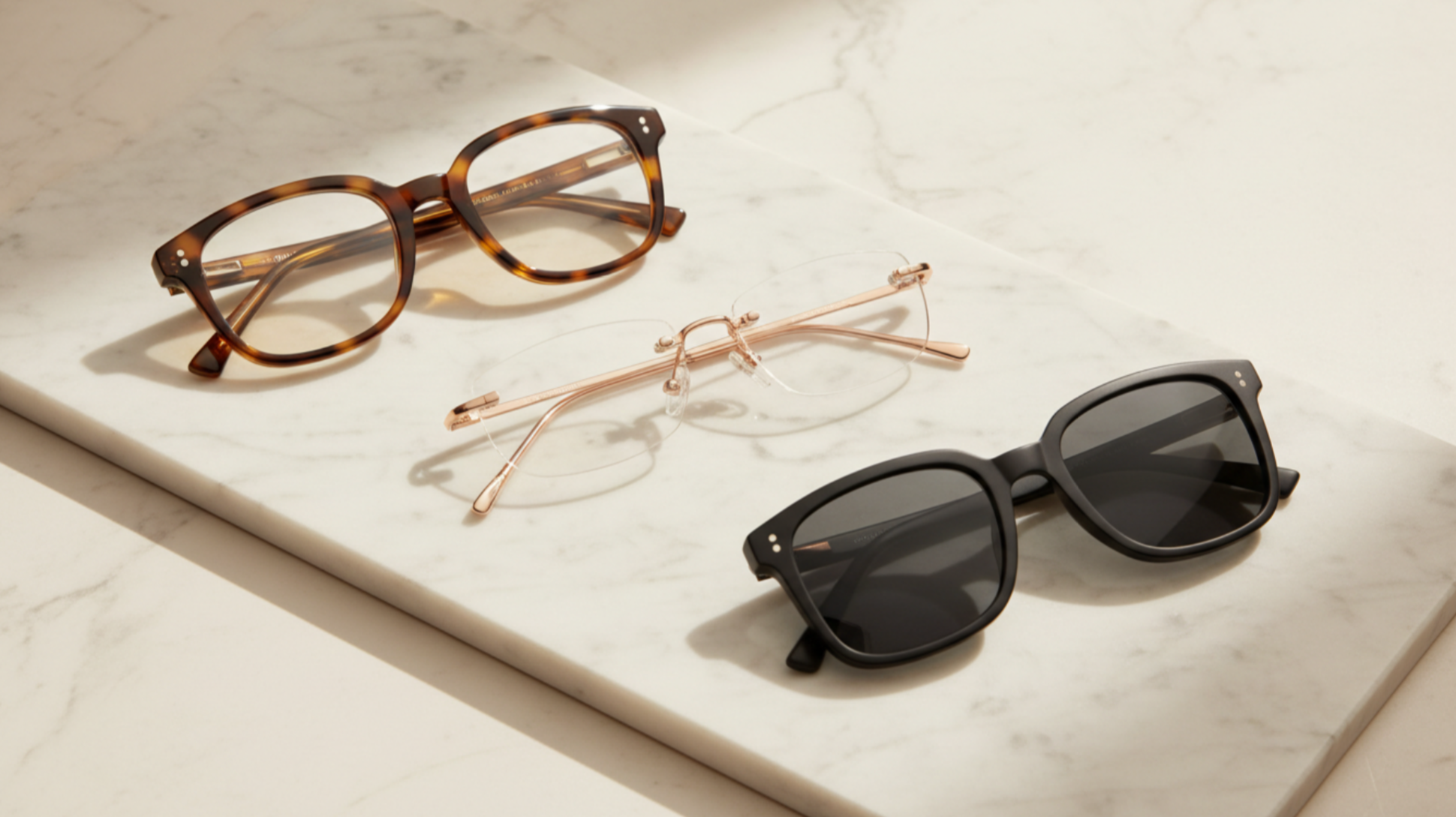 Three pairs of glasses on a white marble surface, including tortoiseshell sunglasses, gold-rimmed reading glasses, and black sunglasses.