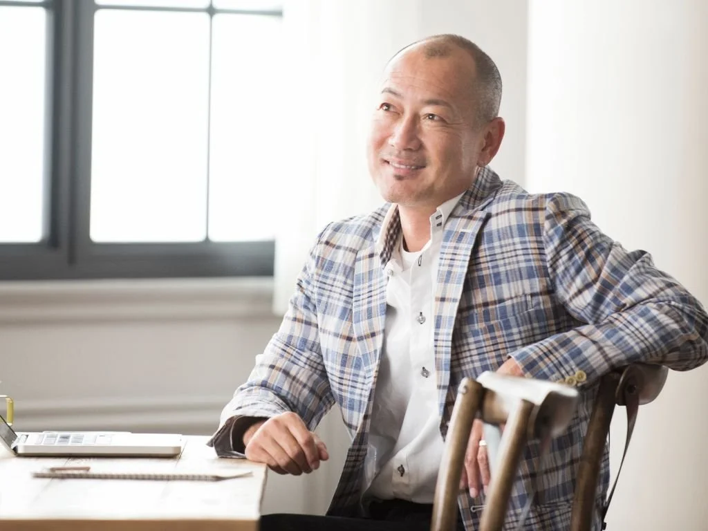 A middle-aged man in a plaid blazer smiling and sitting at a desk in a bright office with large windows.