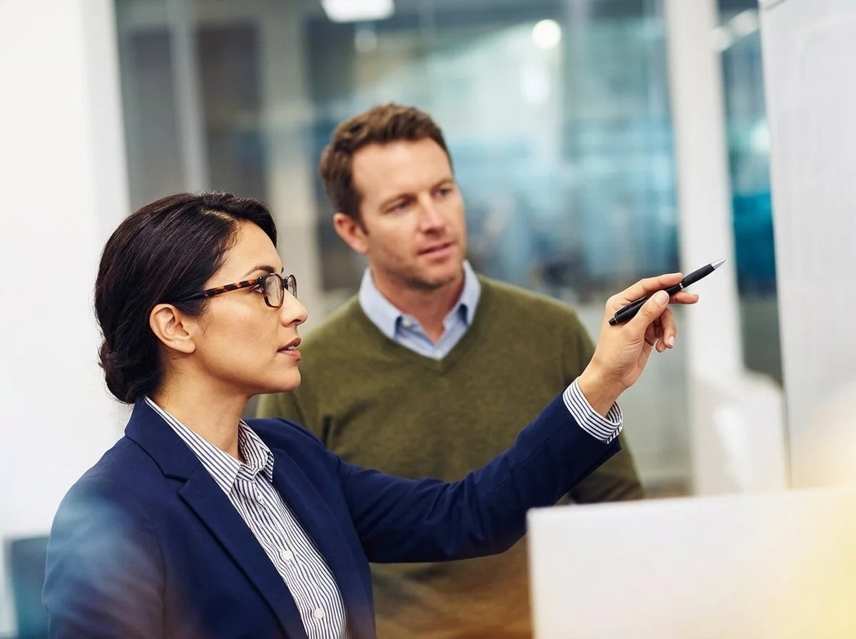 A woman in business attire writing on a whiteboard or presentation screen with a man observing.