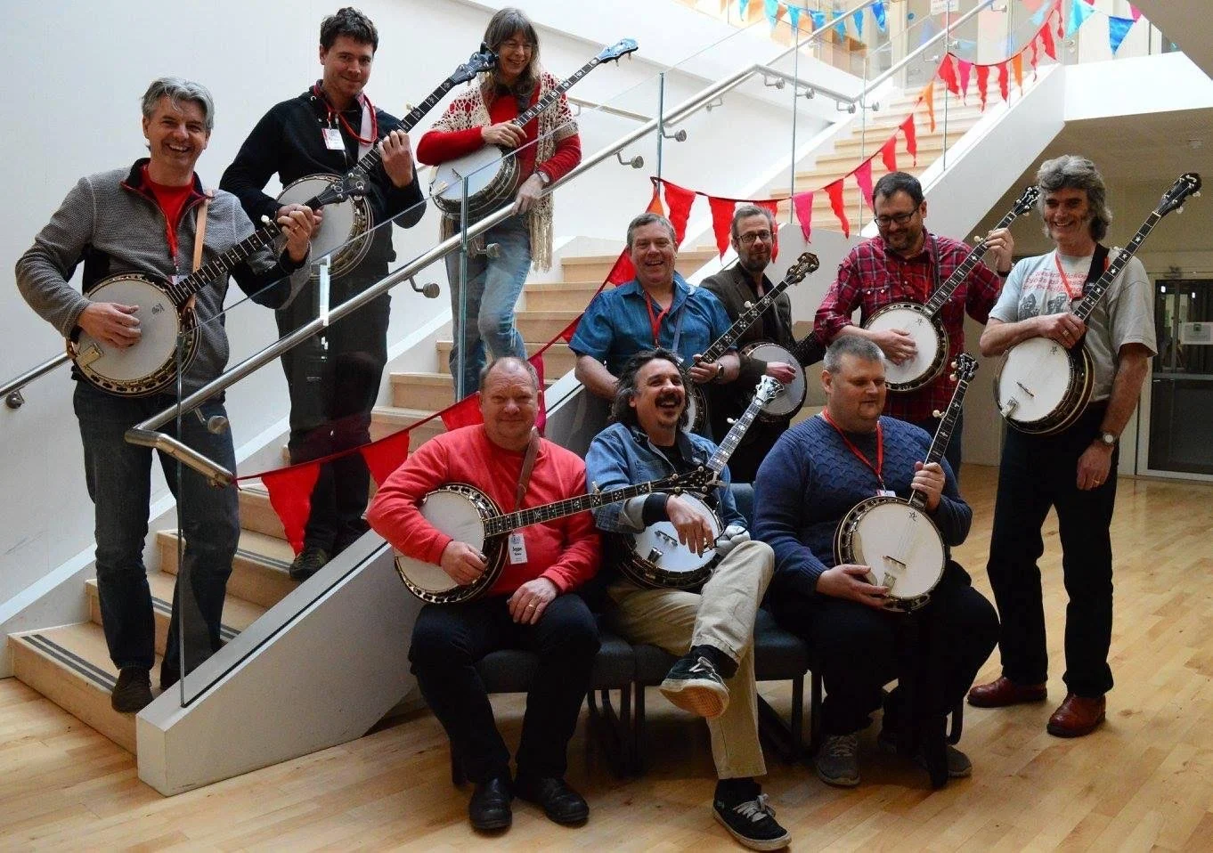 A group of ten people, holding banjos and guitars, posing on a staircase decorated with red and orange flags at an indoor event, smiling and enjoying themselves.