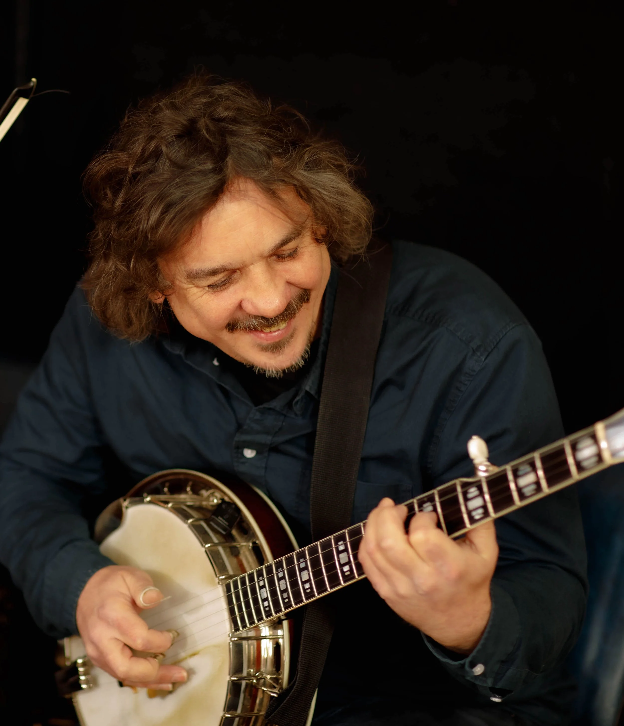 A man with curly brown hair, a beard, and a black shirt playing an acoustic guitar against a black background.