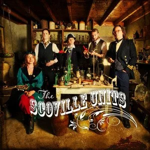 Five people dressed in vintage attire standing around a table with drinks inside a rustic room, with a logo reading 'The Scoville Units' in front.
