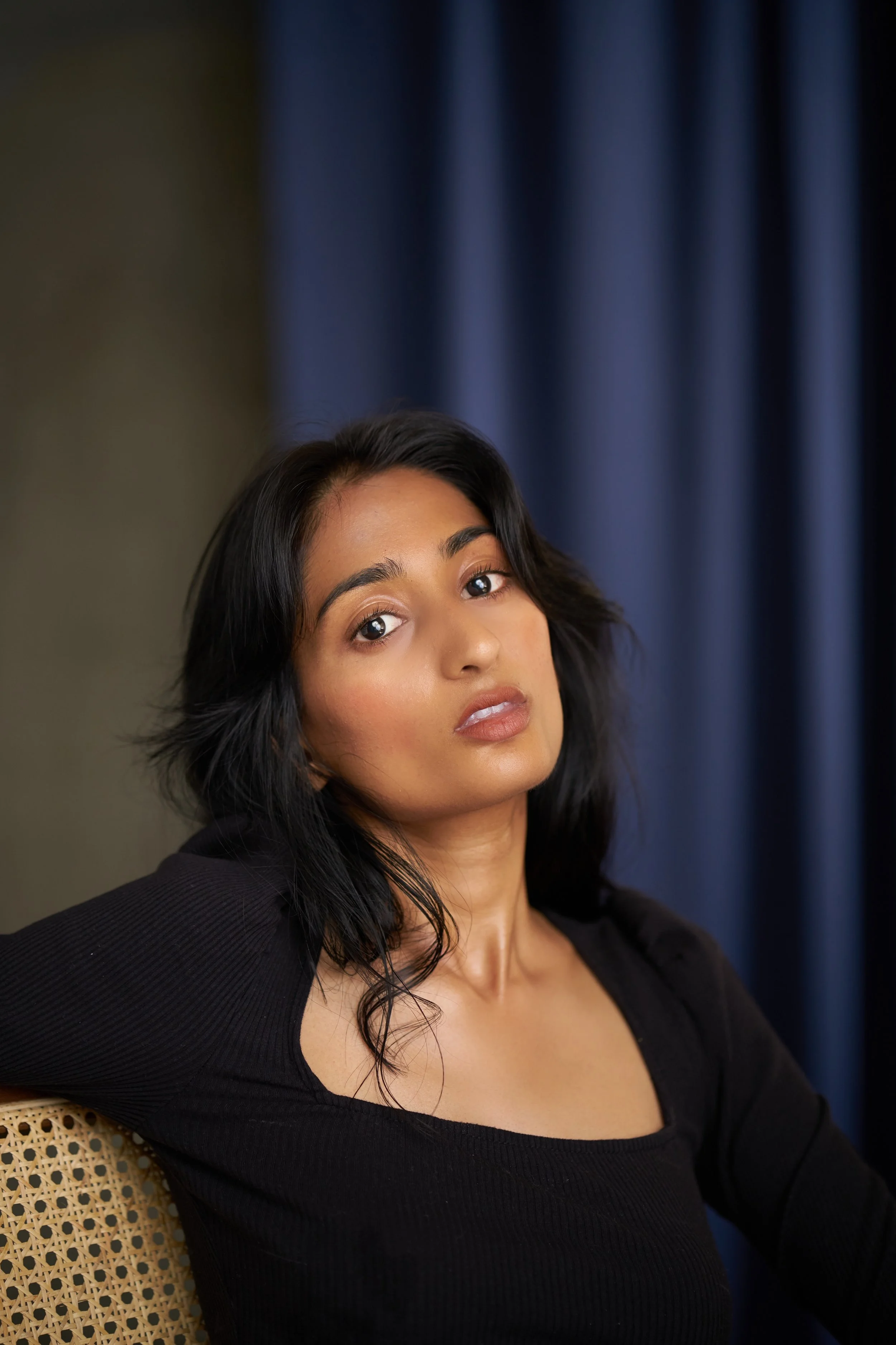 A woman with dark hair, wearing a black top, is sitting on a wicker chair with a patterned back. She is looking at the camera with a neutral expression, and a blue curtain is in the background.