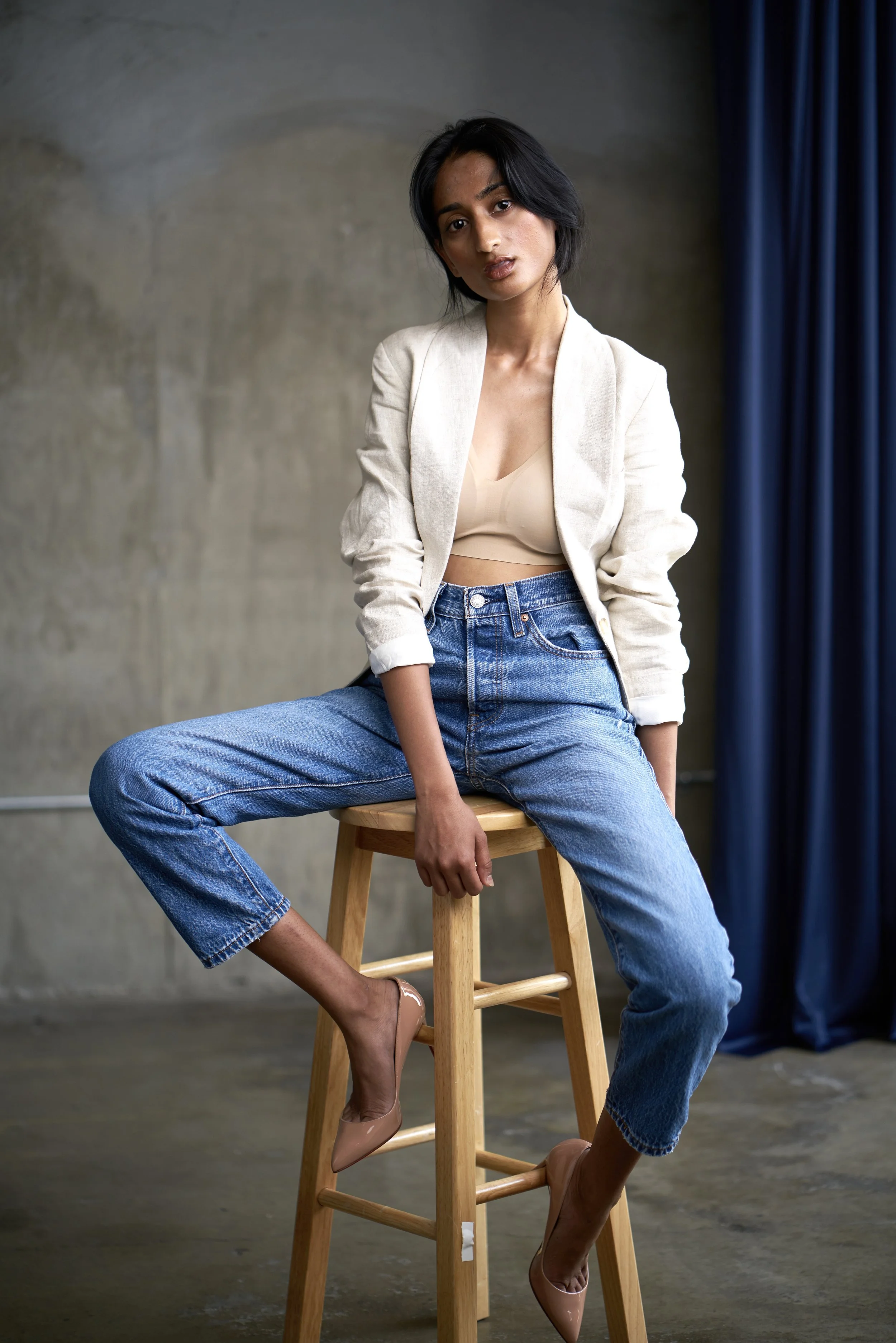 A woman with dark hair and medium skin tone sitting on a wooden stool, wearing a beige crop top, a cream blazer, and high-waisted blue jeans, with one leg crossed over the other, in front of a concrete wall and dark blue curtains.