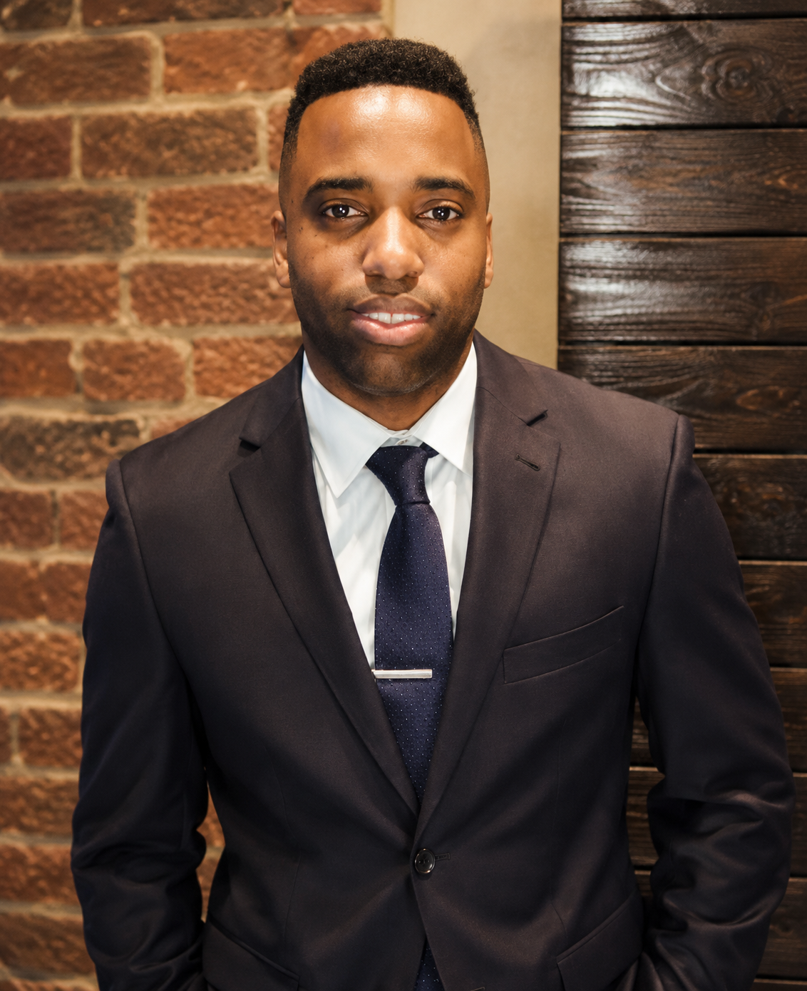 A man dressed in a black suit with a white shirt and dark tie, standing against a background of brick and wood wall. He is looking at the camera with a slight smile.