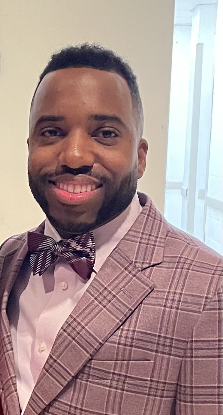A smiling man with a well-groomed beard wearing a checkered brown suit, white shirt, and a striped bow tie, standing indoors near a window.