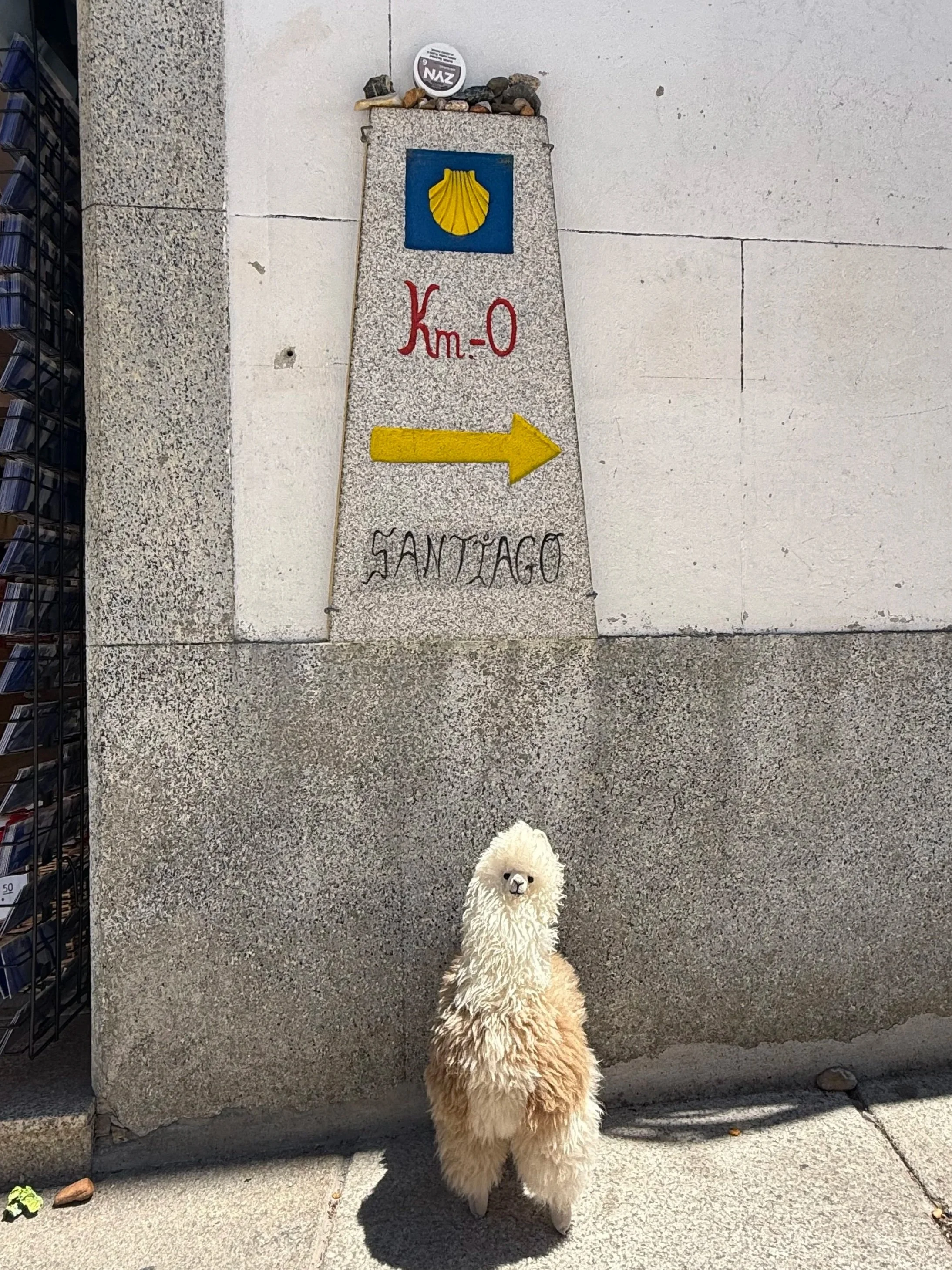 A plush llama toy standing on a sidewalk in front of a stone wall with a sign that reads 'Km-0' and a yellow arrow pointing to the right. The sign also has a blue square with a yellow shell and some rocks on top.