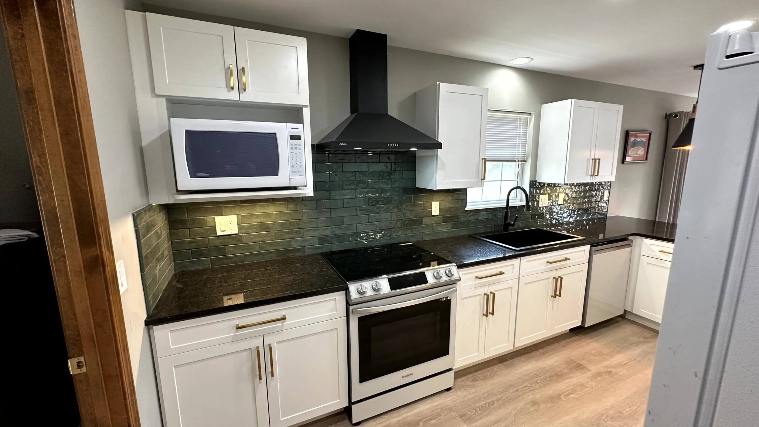 A modern kitchen with white cabinets, black countertops, a black range hood, and a black stovetop. The backsplash consists of dark green subway tiles. There are a microwave and a white refrigerator visible, along with a window above the sink.