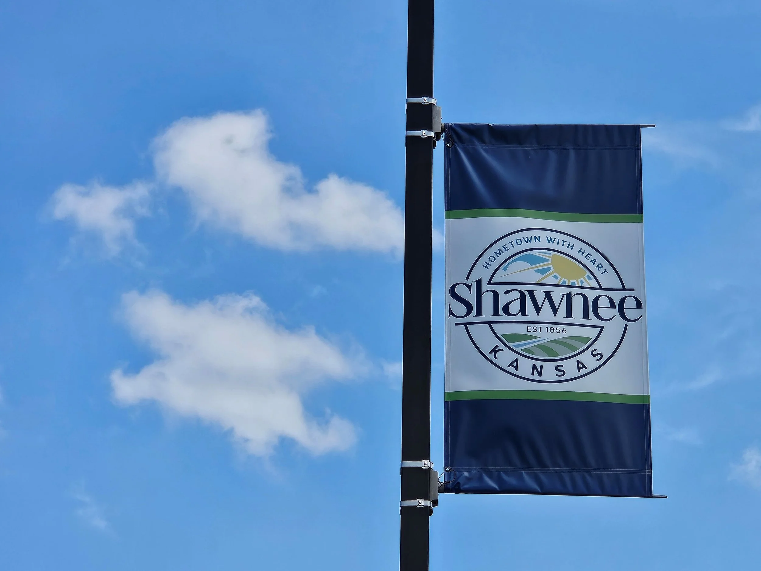 A flag with a circular emblem reading 'Hometown with Heart Shawnee Kansas' and the year 1856, mounted on a pole against a blue sky with clouds.