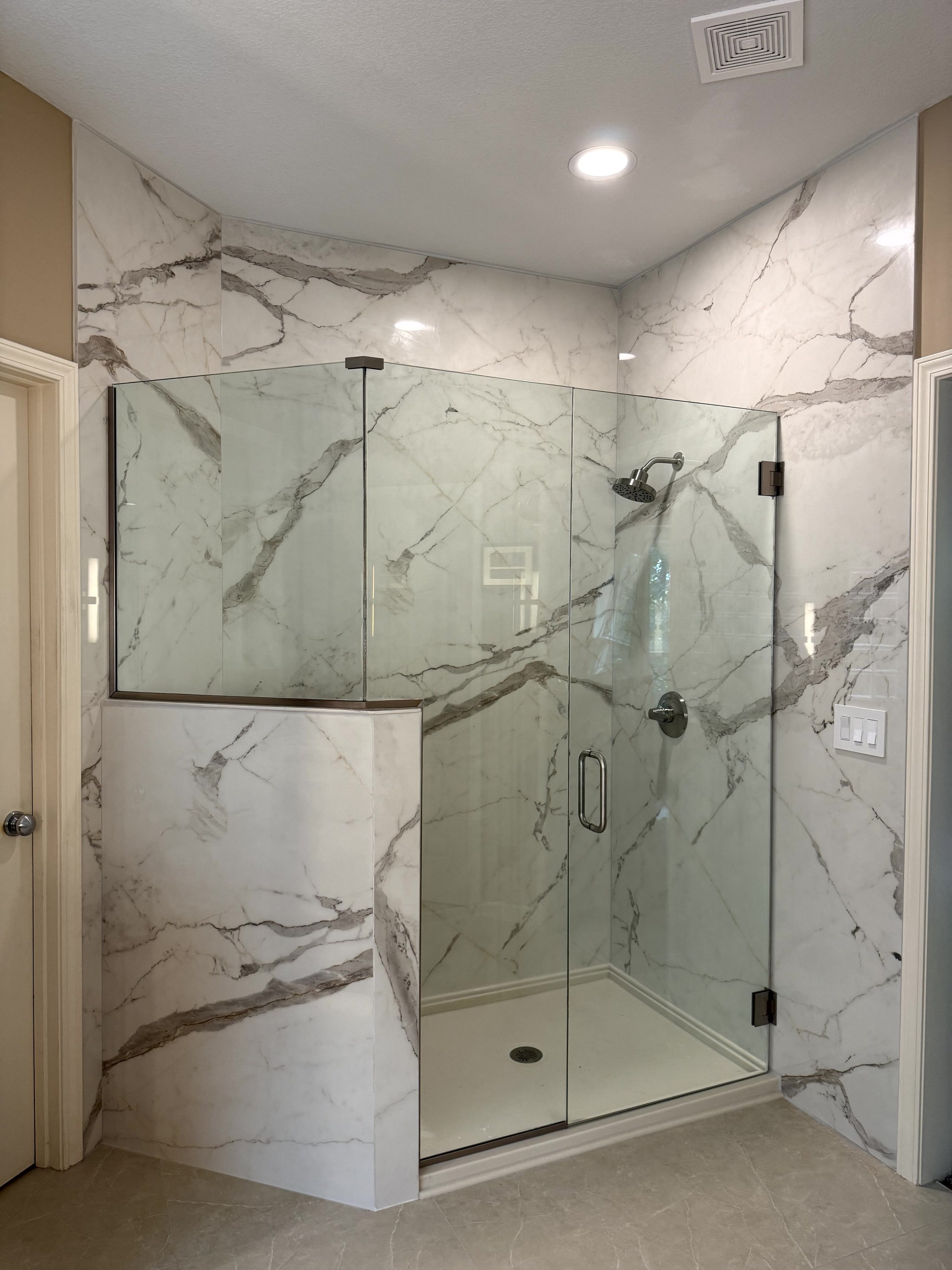 A modern walk-in shower with glass enclosure, white marble walls with gray veins, a silver showerhead, and a built-in shelf, with beige and cream-colored tiles on the floor and surrounding walls.