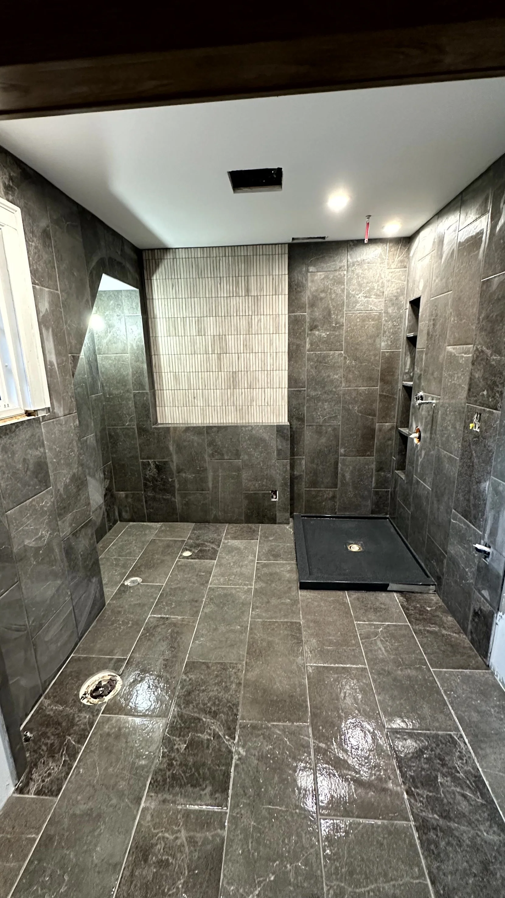 Bathroom under construction with dark gray tile flooring and walls, a small window, recessed shelves, and an empty shower base.