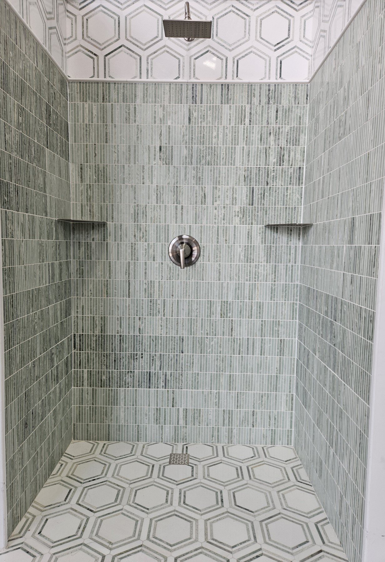 A modern shower with gray stone tiles on the walls, a mounted square rainfall showerhead, a handle on the wall, and decorative hexagonal tiles on the floor and ceiling.