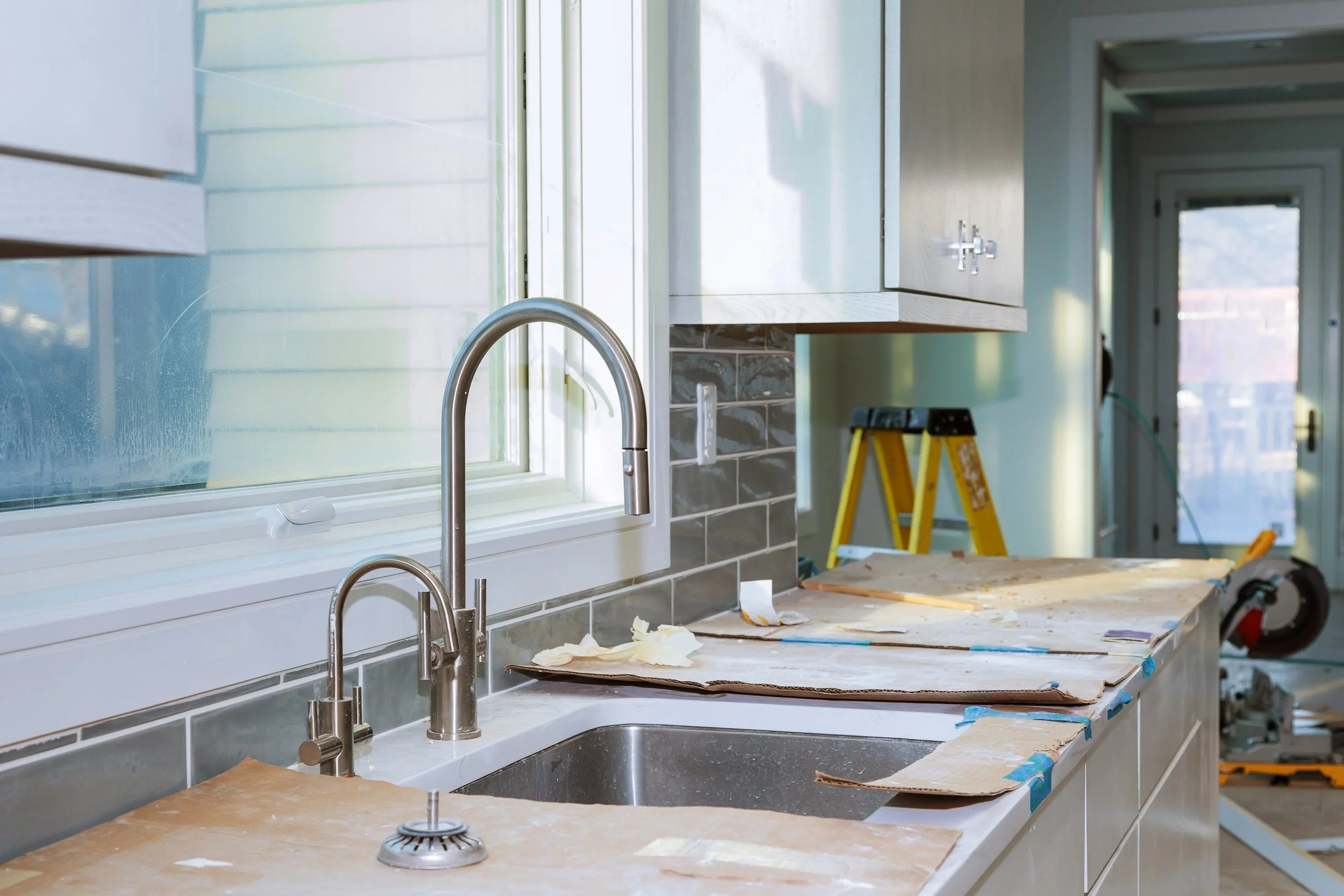 Kitchen counter with construction materials, a window, a faucet, and a yellow step ladder in the background.