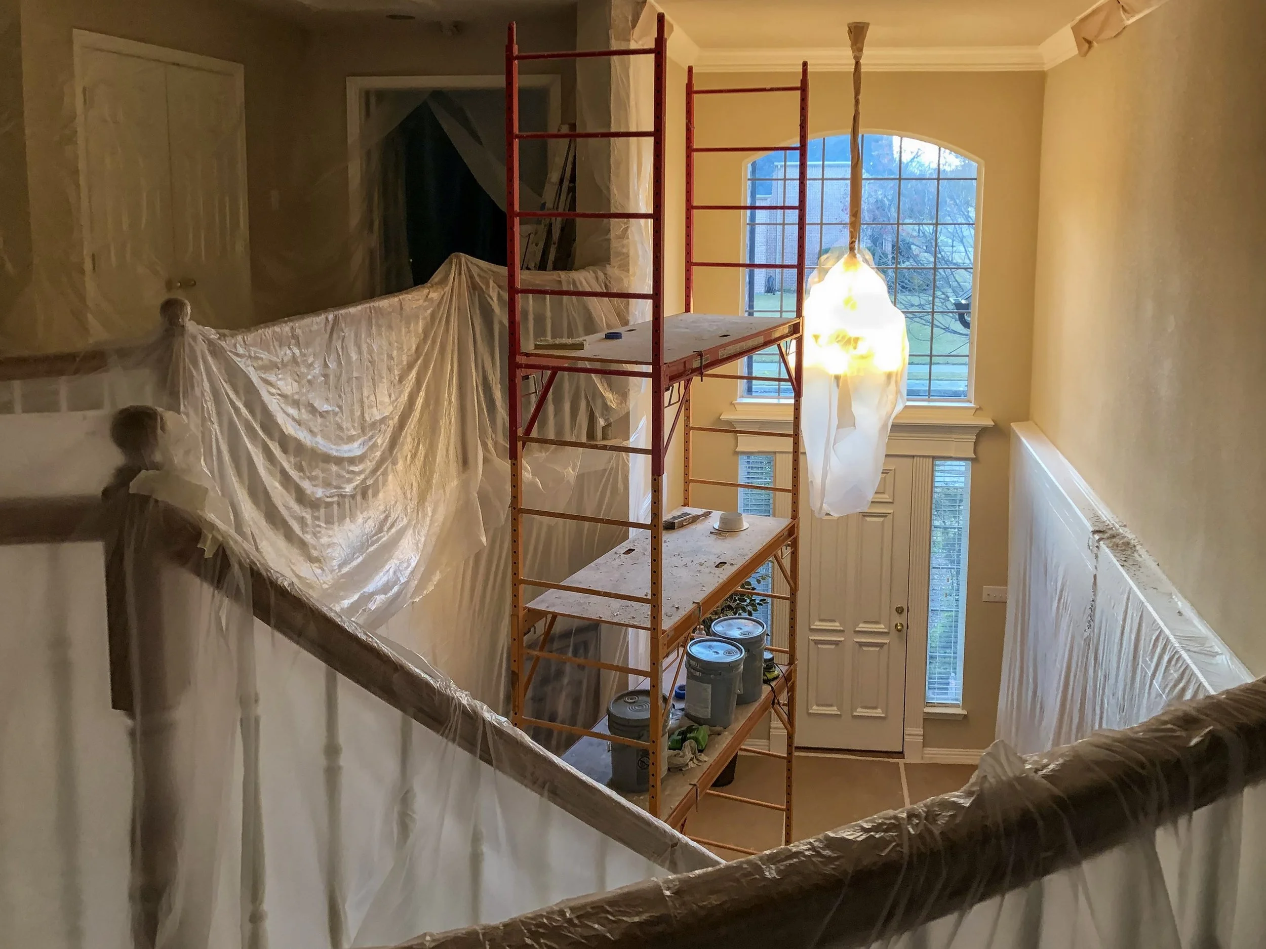 Interior of a house under renovation with scaffolding set up near a large window, covered in plastic sheets, with some paint cans and tools on the floor.