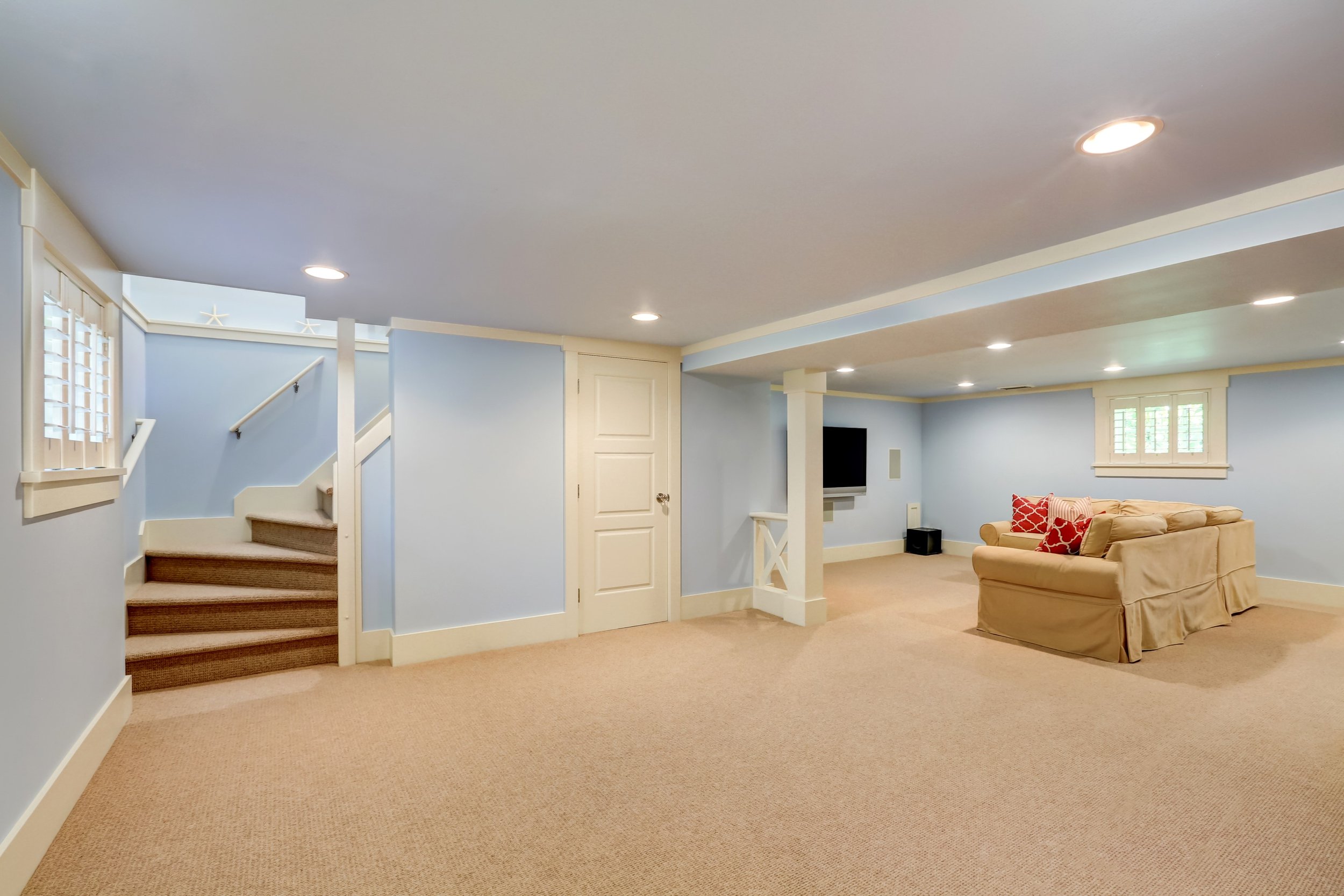 A living room with beige carpet, light blue walls, and white trim. There are recessed ceiling lights, a window with shutters, a staircase leading upstairs, and a doorway. Beige sofa with red and white patterned pillows is on the right side.