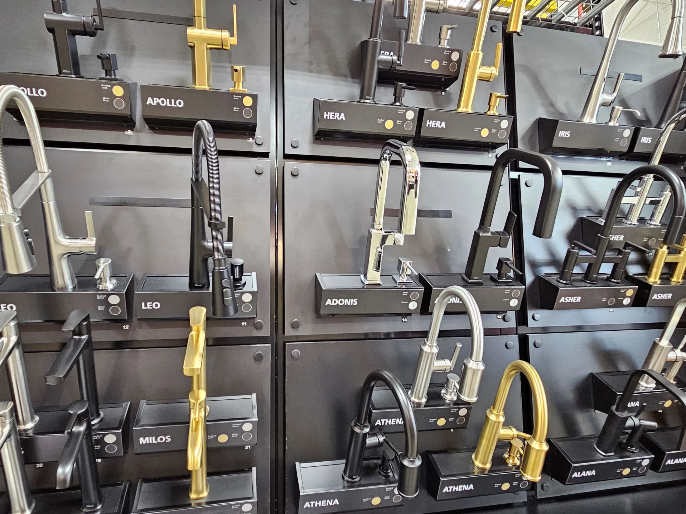 Display of modern kitchen faucets in black and gold colors, labeled with names like Apollo, Hera, Leo, Athena, Alana, and Iris, arranged on black shelves.