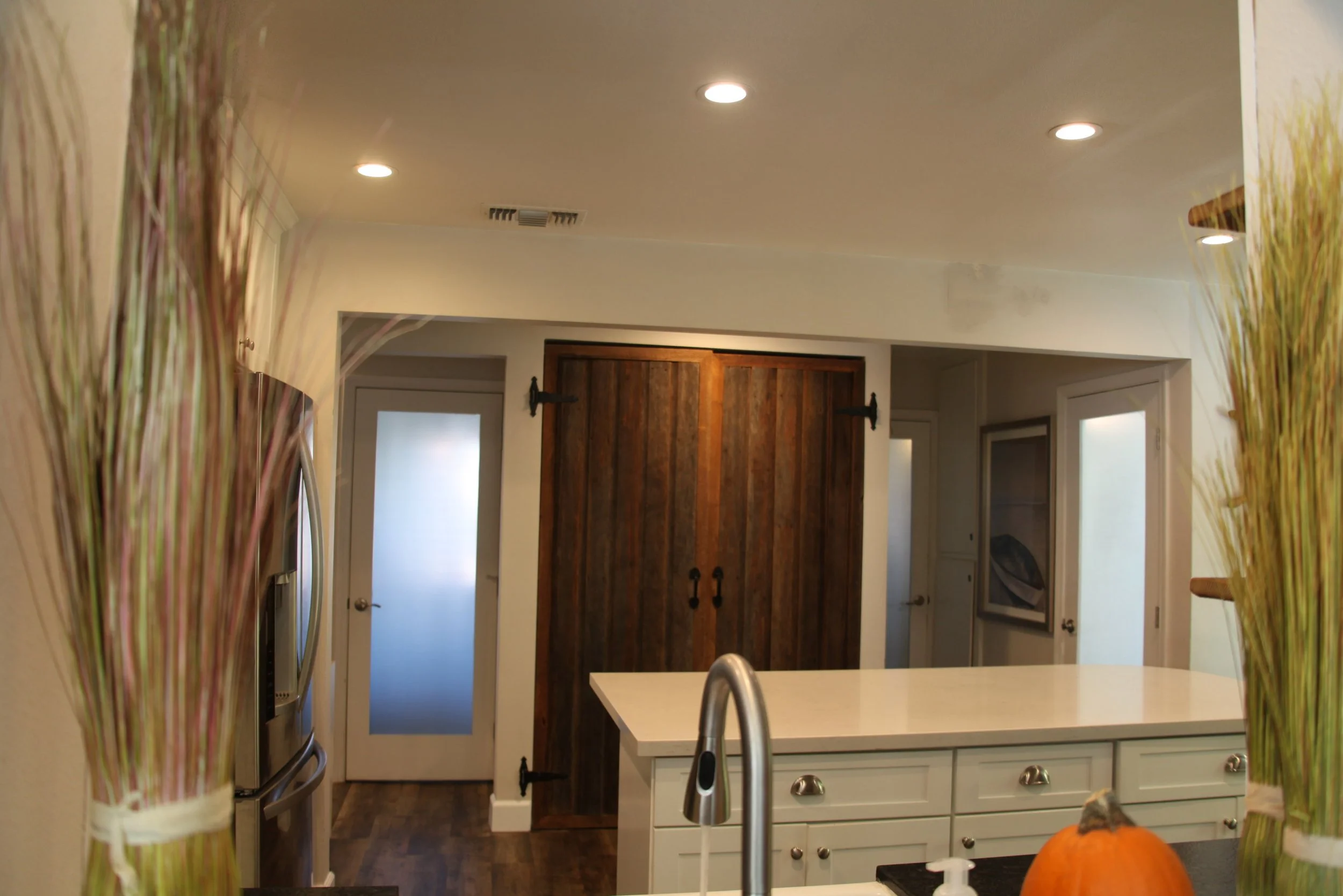 A kitchen view looking towards a doorway with a wooden sliding barn door, a white kitchen island with a sink, and decorative plants with pumpkins in the foreground. Recessed ceiling lights illuminate the space.