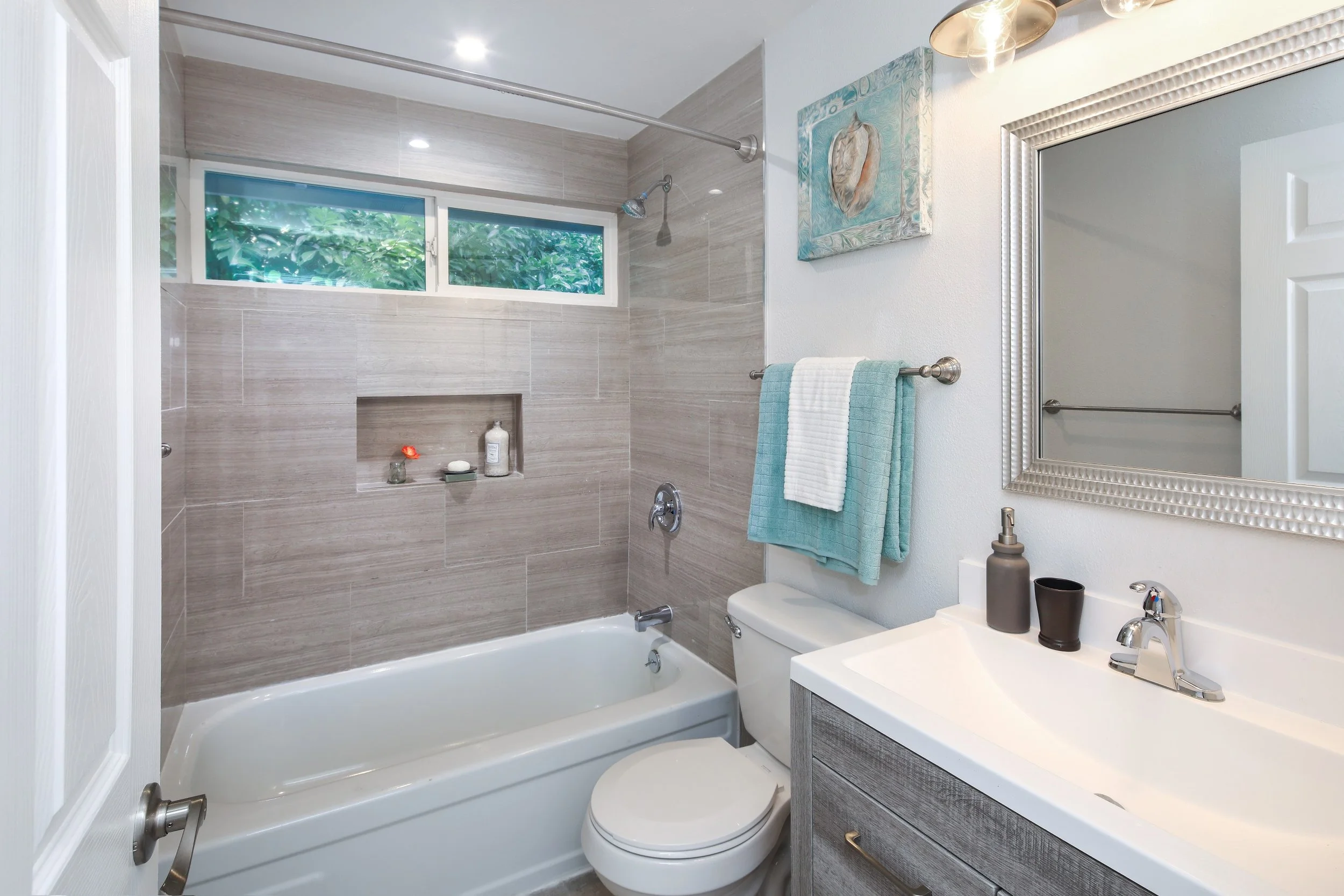 A modern bathroom with beige tiled walls, a white bathtub, a toilet, a sink with a gray cabinet, a mirror with a beaded silver frame, a towel bar with a turquoise and white towel, a window with greenery outside, and artistic wall decor.