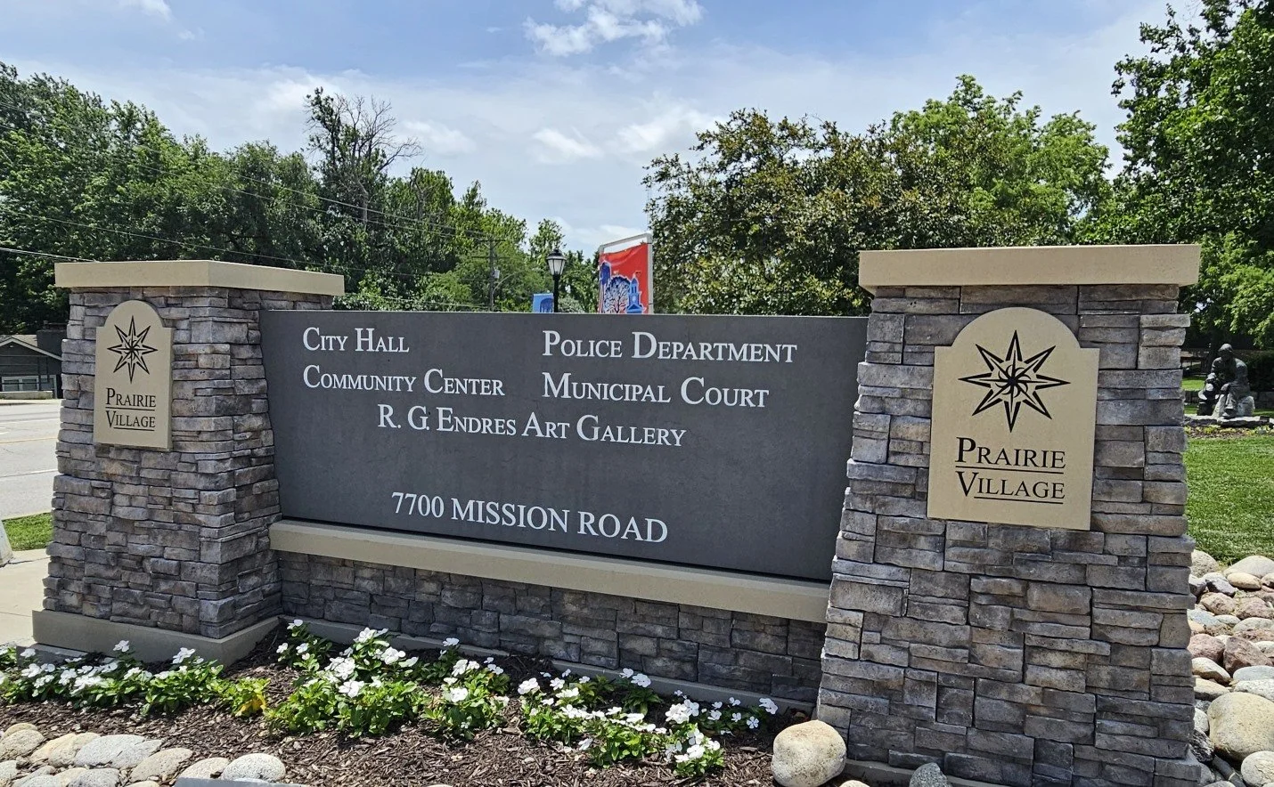 Sign for Prairie Village at 7700 Mission Road, indicating city hall, police department, community center, municipal court, and art gallery, with decorative brick columns and flower bed in foreground.