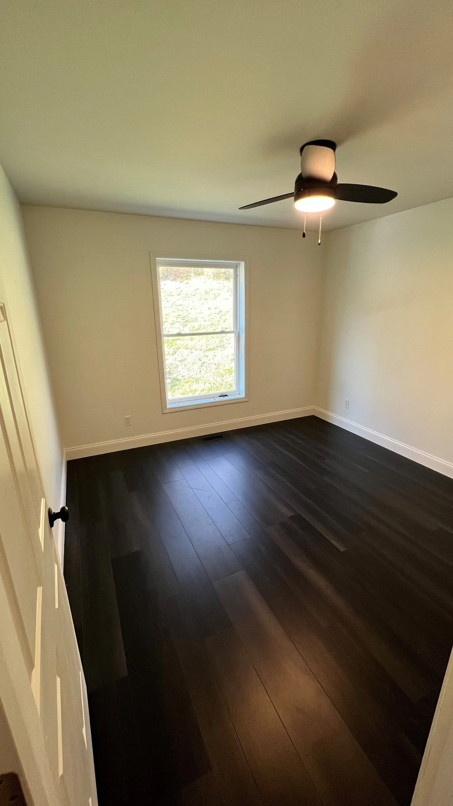 Empty room with dark hardwood floors, white walls, a single window, and a ceiling fan with a light.